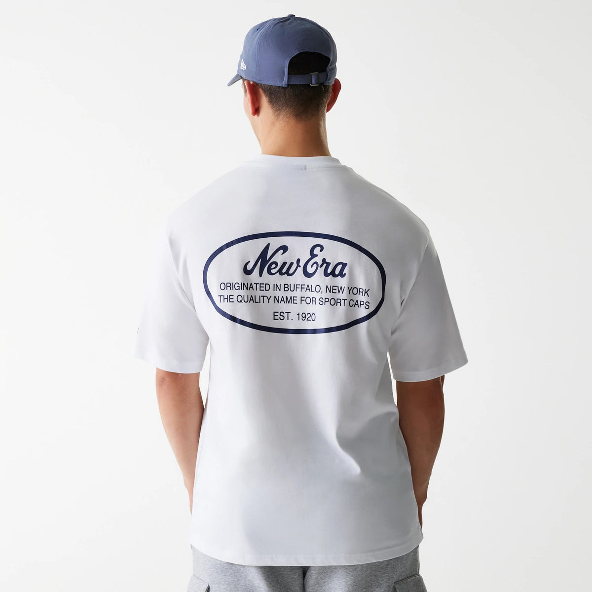 New Era Trucker Patch White Oversized T-Shirt, rear view showing a large blue printed oval logo centred on the upper back with text, worn by a person in grey shorts with a debossed New Era logo on a blue cap.