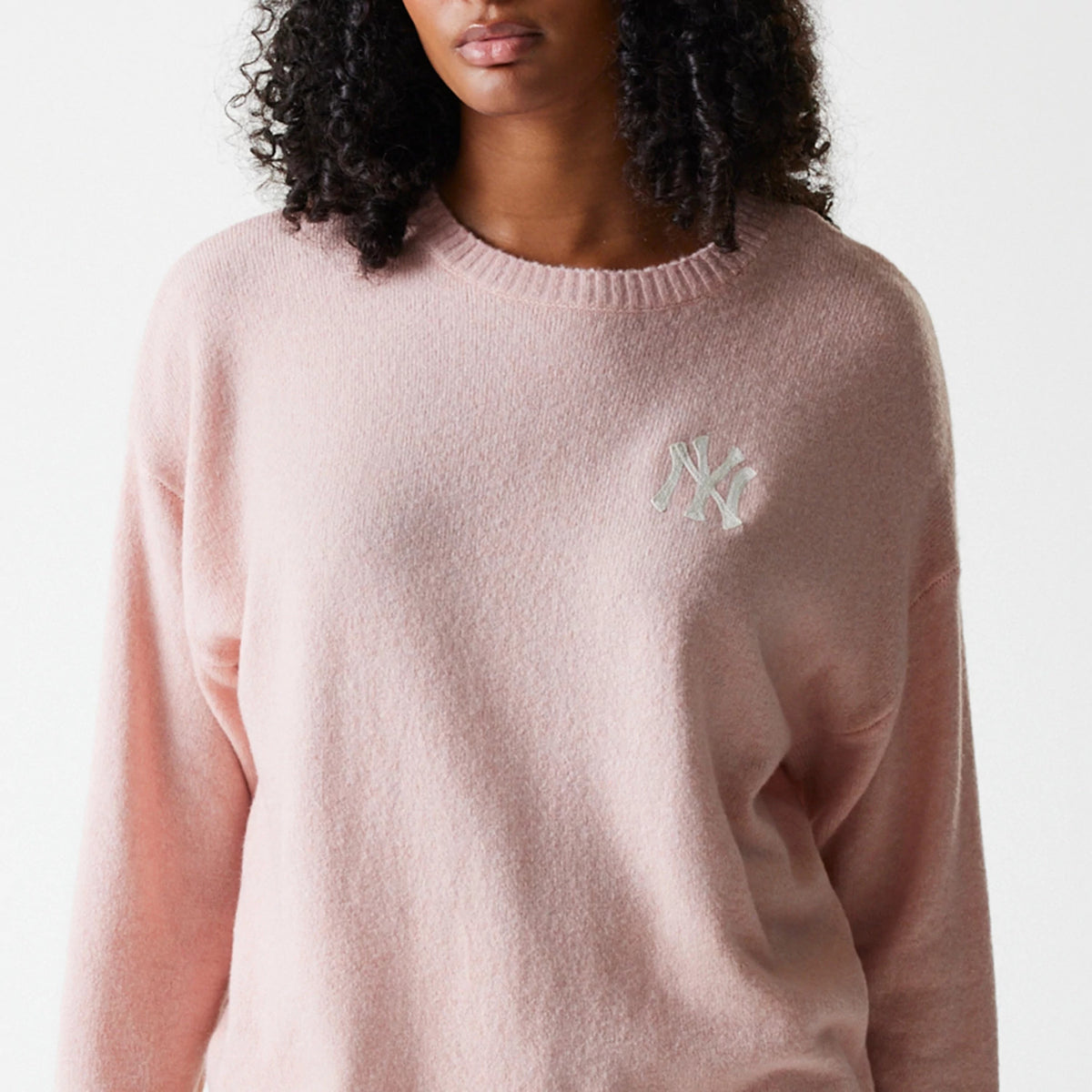 New Era New York Yankees Womens MLB Knit Pastel Pink Crew Neck Sweater, front view on a model with curly hair, showcasing the white embroidered NY logo on the left chest of the soft pastel pink fabric.