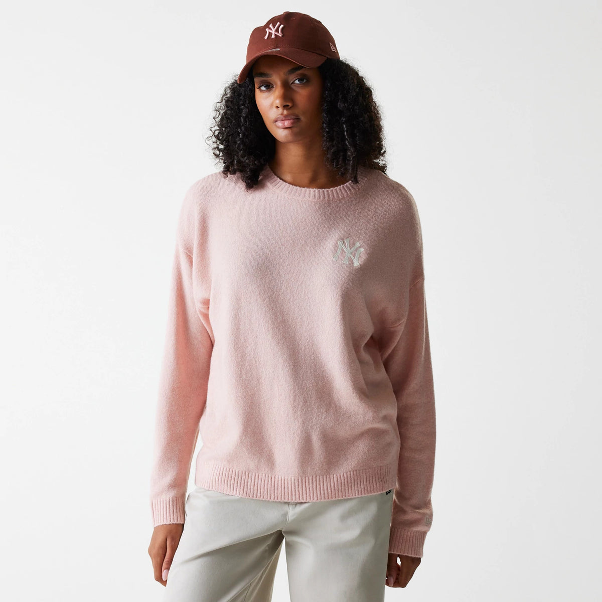 New Era New York Yankees Womens MLB Knit Pastel Pink Crew Neck Sweater, on-model view styled with a brown cap, showing the white embroidered NY logo on the left chest of the soft pastel pink sweater.