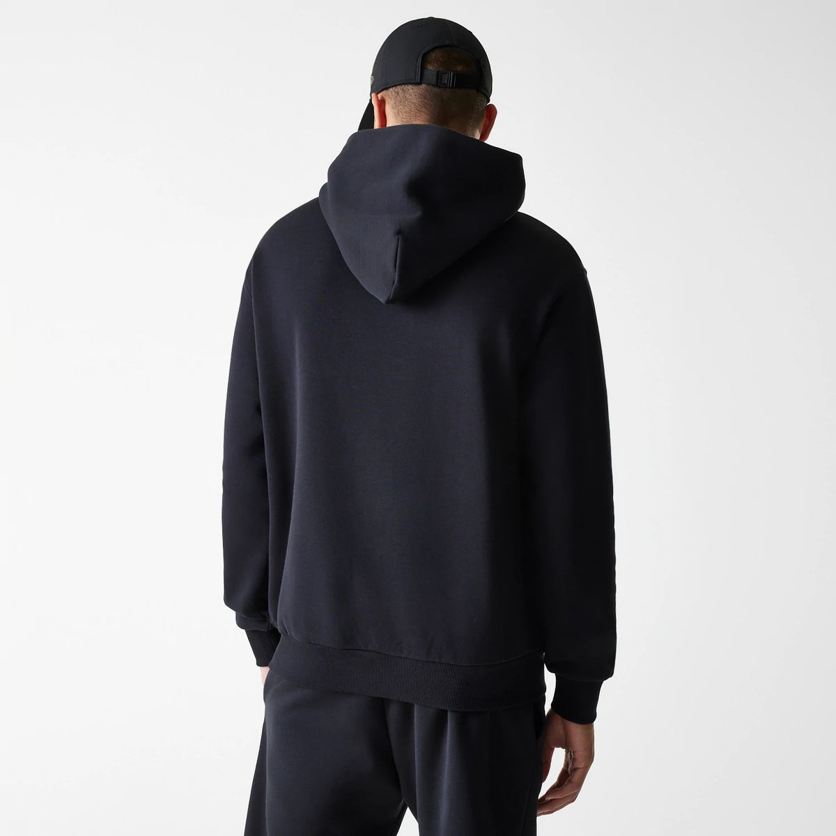 New Era Tech Black on Black Oversized Pullover Hoodie, rear view showing the relaxed fit with a large hood draping down, ribbed cuffs and hem, and a cap with a debossed New Era logo worn by the model.