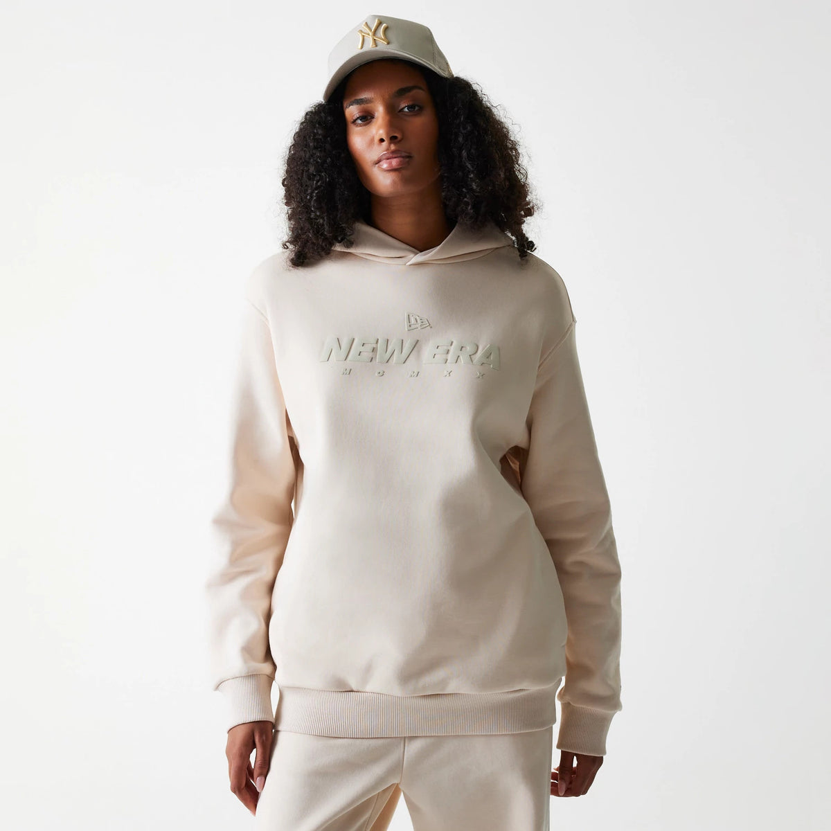 New Era Tech Light Beige Oversized Pullover Hoodie, front view showing the embossed New Era logo prominently across the chest, features a loose fit with ribbed cuffs and hem, worn by a model also wearing a matching beige cap.