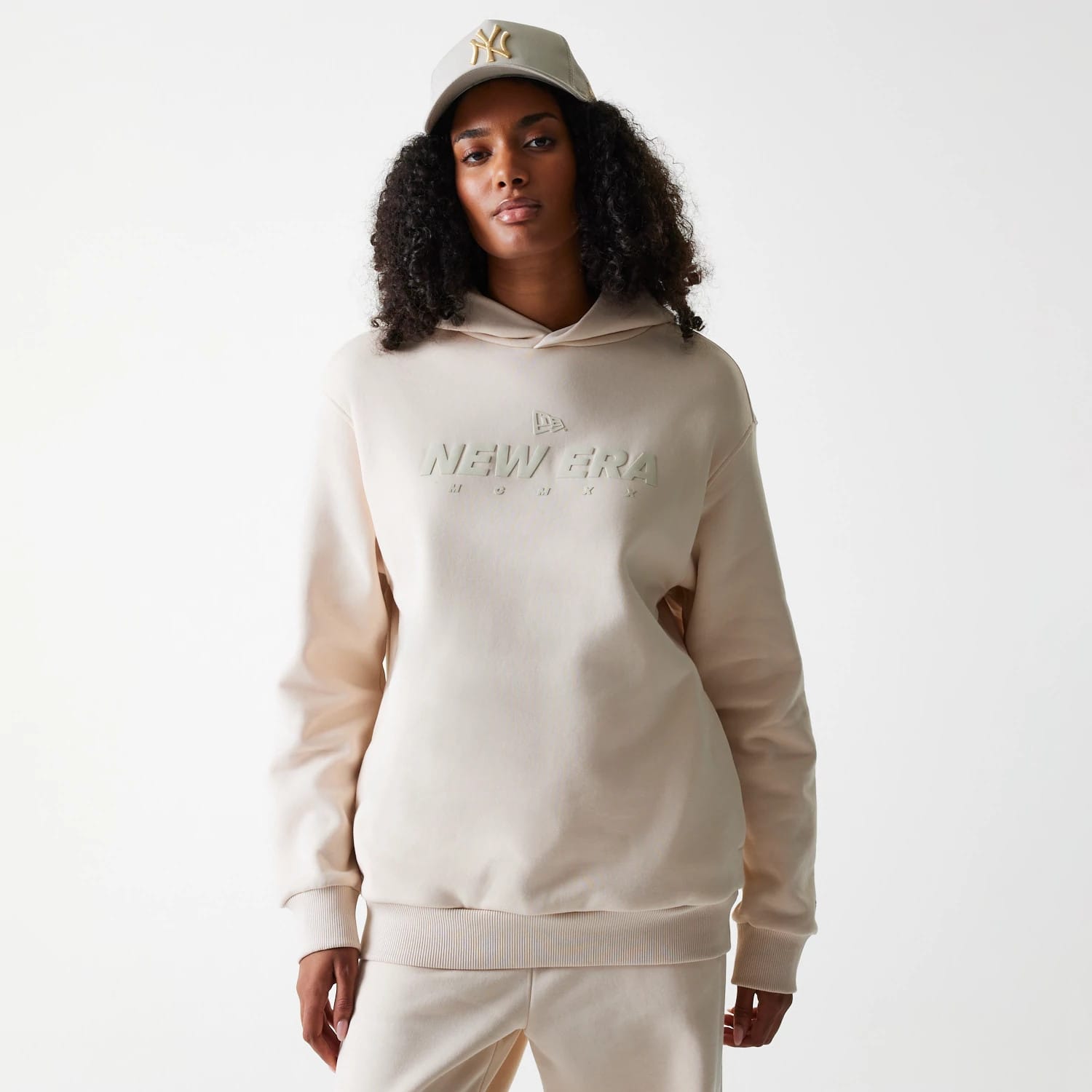 New Era Tech Light Beige Oversized Pullover Hoodie, front view showing the embossed New Era logo prominently across the chest, features a loose fit with ribbed cuffs and hem, worn by a model also wearing a matching beige cap.
