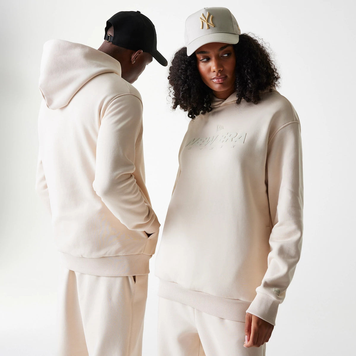 New Era Tech Light Beige Oversized Pullover Hoodie, front view showing the oversized beige hoodie with a debossed New Era logo on the chest, ribbed cuffs, and hem, paired with matching trousers and a cream cap featuring an embossed logo on the front.