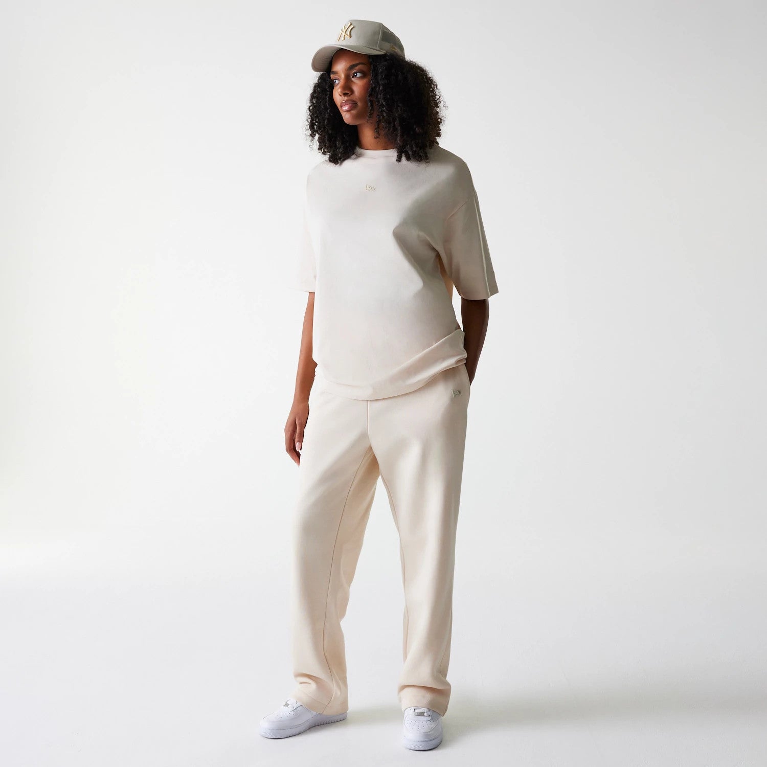New Era Light Beige Straight Leg Joggers, three-quarter view on a model styled with a matching light beige T-shirt and cap, featuring a relaxed fit and subtle logo detail on the left leg.