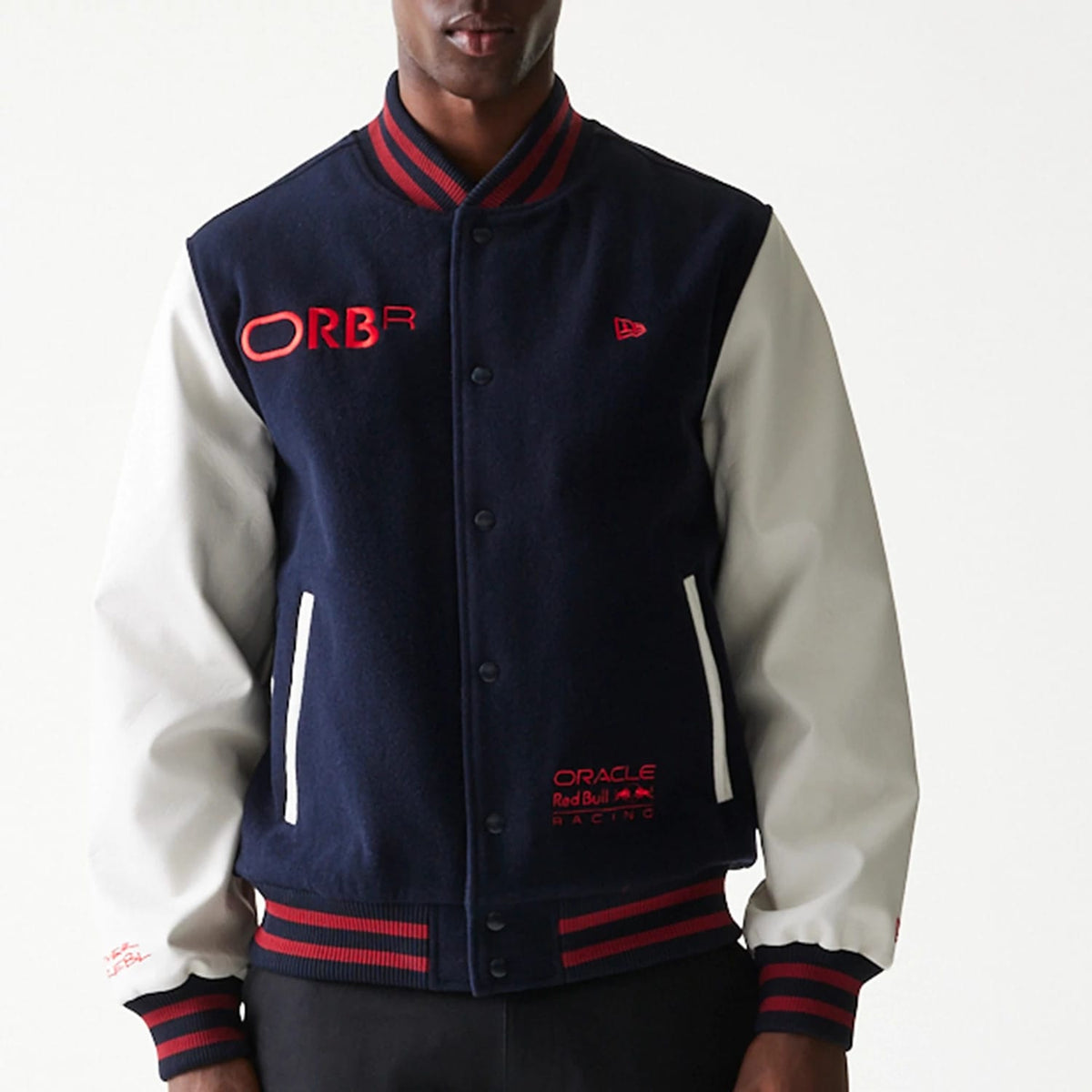 New Era Red Bull Racing Dark Blue Varsity Jacket, front view showing the dark blue body with white sleeves, embroidered ORBR logo in red on the chest, Oracle Red Bull Racing logo at the bottom, and striped red and blue collar and cuffs.