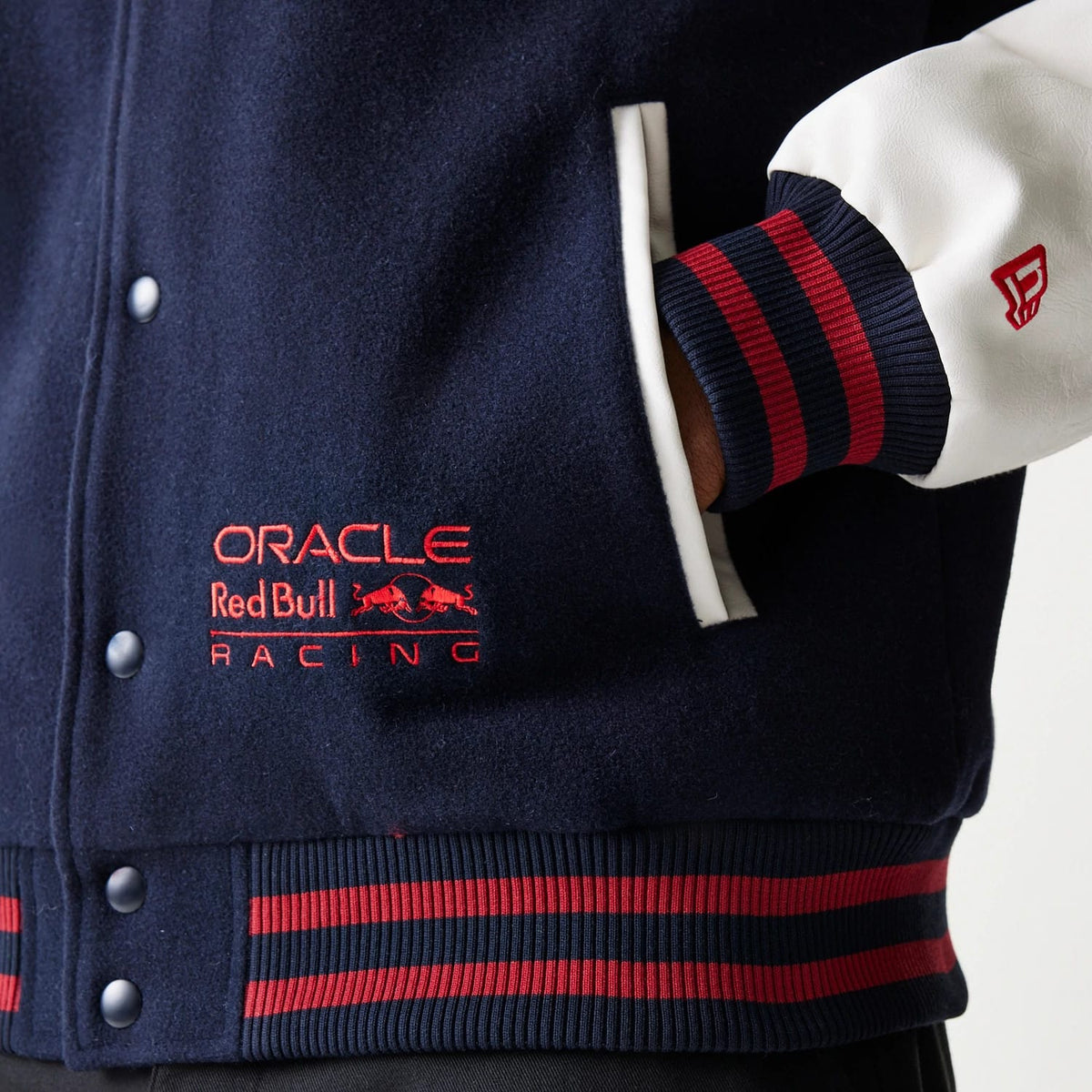 New Era Red Bull Racing Dark Blue Varsity Jacket, three-quarter view showing the dark blue fabric with red embroidered logo on the chest, white sleeves with red and navy striped cuffs, and snap button closure along the front.