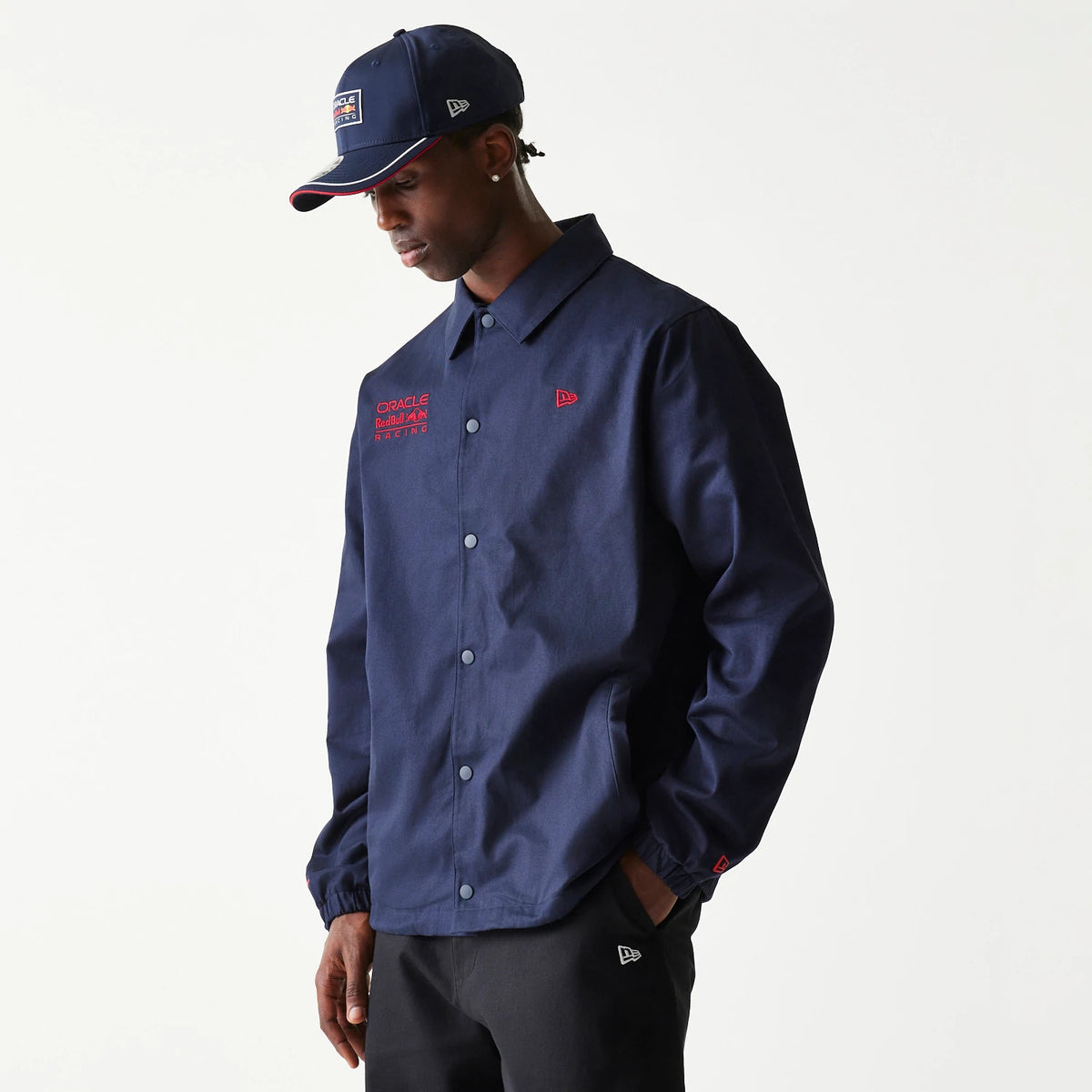 New Era Red Bull Racing Dark Blue Oversized Coach Jacket, on-model view featuring a dark blue jacket with red embroidered logos on the chest, a button-up front, and a matching cap with a raised New Era logo on the side.