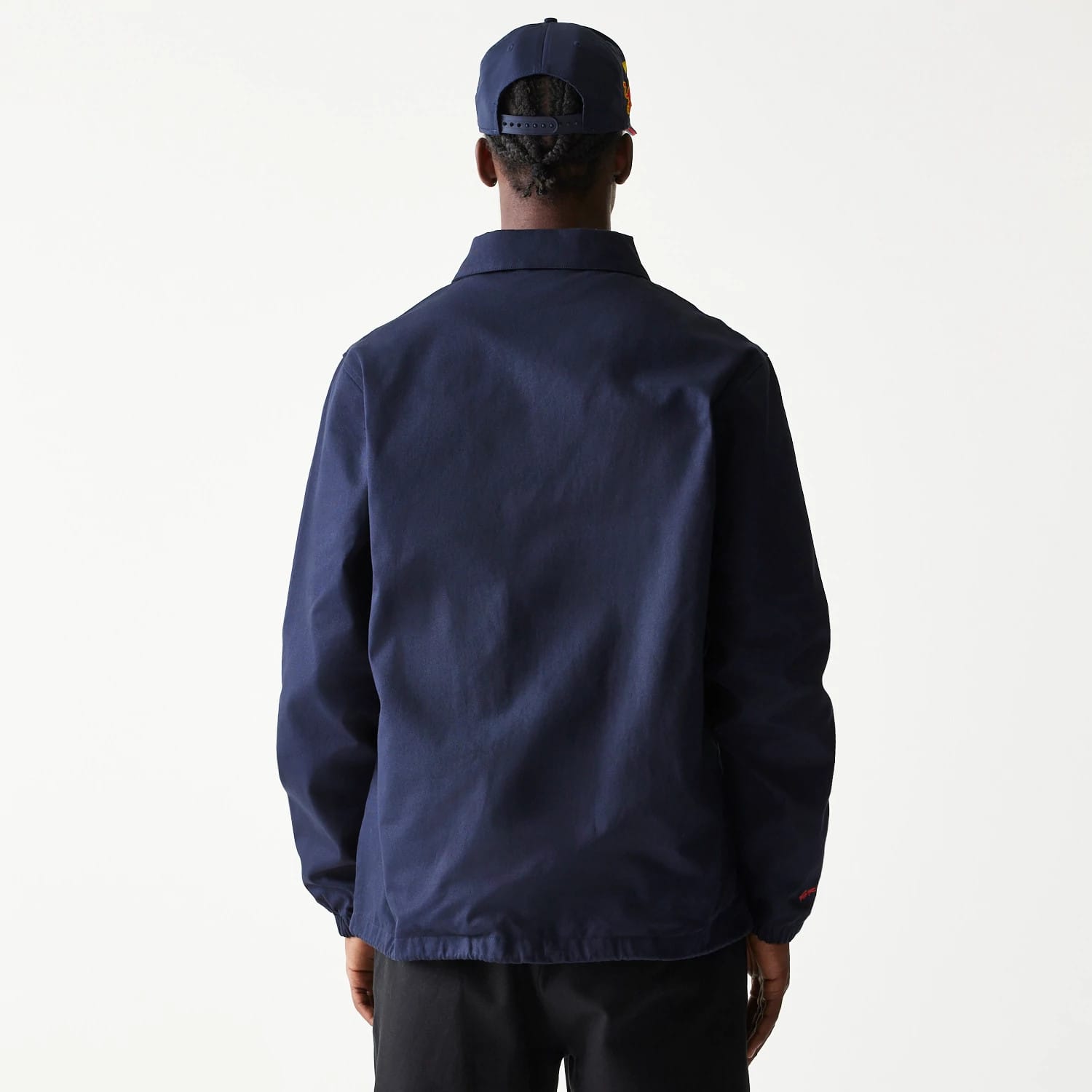 New Era Red Bull Racing Dark Blue Oversized Coach Jacket, rear view showing the plain dark blue back with a relaxed fit, long sleeves, and a small debossed logo on the lower right sleeve cuff, worn by a model in a matching dark blue cap.
