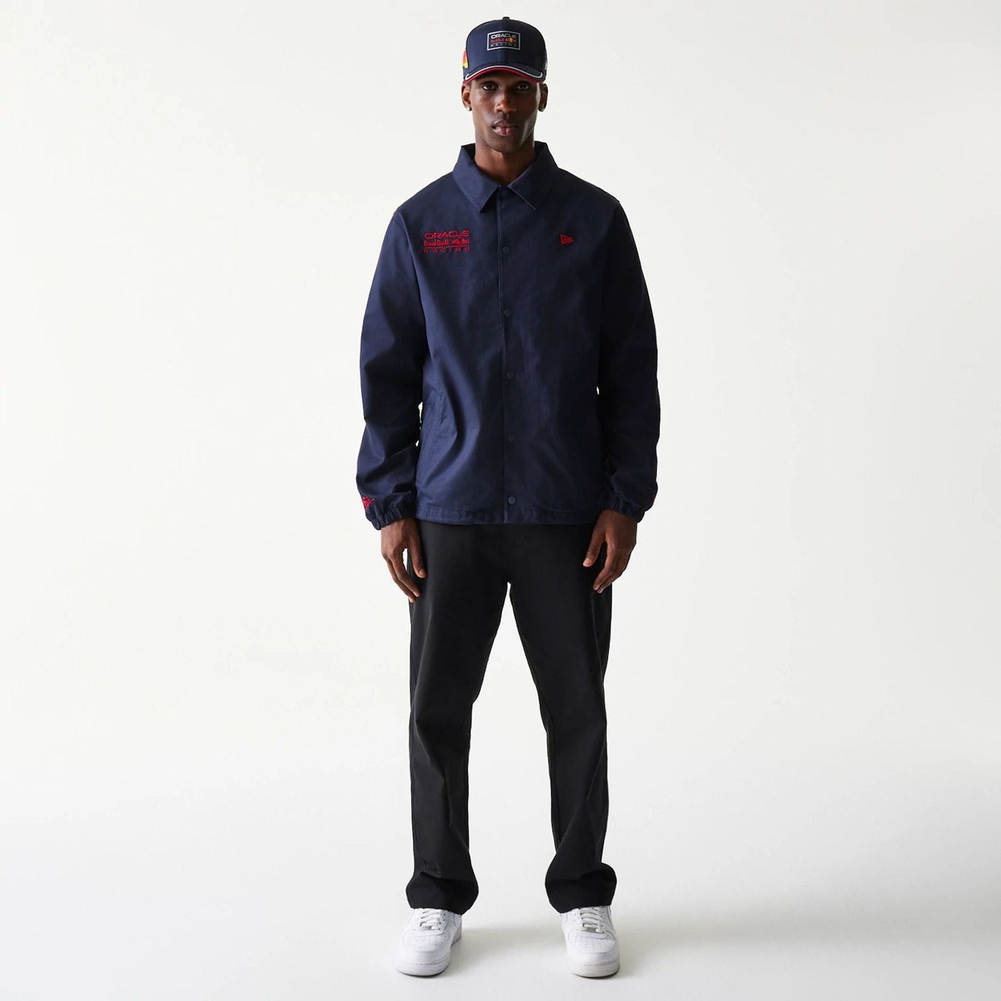 New Era Red Bull Racing Dark Blue Oversized Coach Jacket, on-model view showing a dark blue jacket with a button-down front, red logo on the chest, and matching cap worn by the model, styled with black trousers and white trainers.
