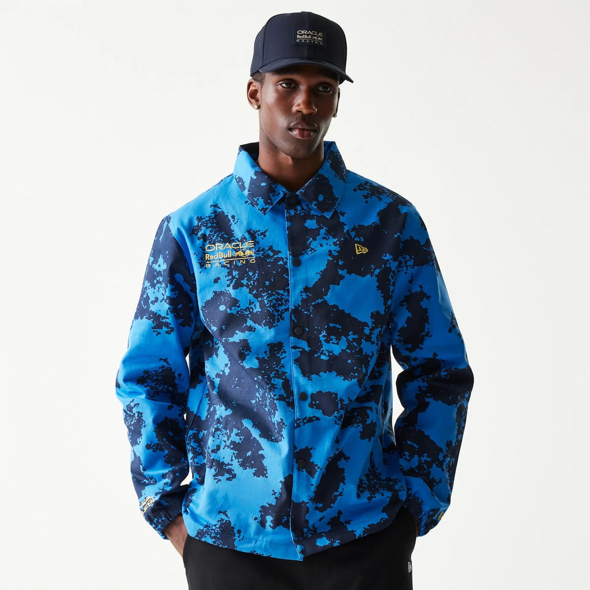 New Era Red Bull Racing Blue Oversized Coach Jacket, front view showing a blue and black patterned design with a debossed New Era logo on the left chest, the Red Bull Racing logo in yellow on the right chest, and button-down closure.