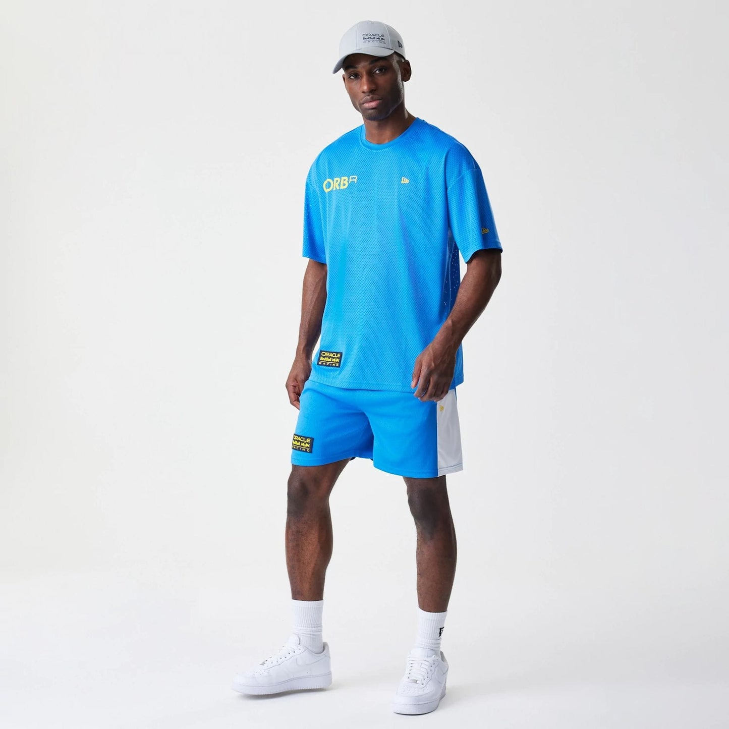New Era Red Bull Racing Graphic Mesh Blue Shorts, on-model view highlighting the blue colour with white side panels, featuring a printed logo on the side, styled with a coordinating blue top and white trainers.