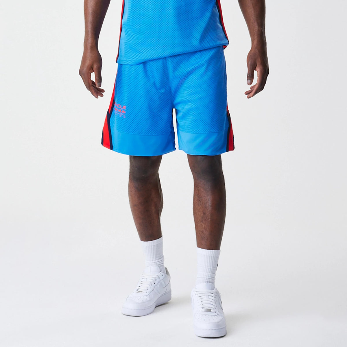New Era Red Bull Racing Basketball Mesh Blue Shorts, front view on-model showing the bright blue mesh fabric with red panels on the sides, an embroidered Red Bull Racing logo on the left leg, paired with white trainers and socks.