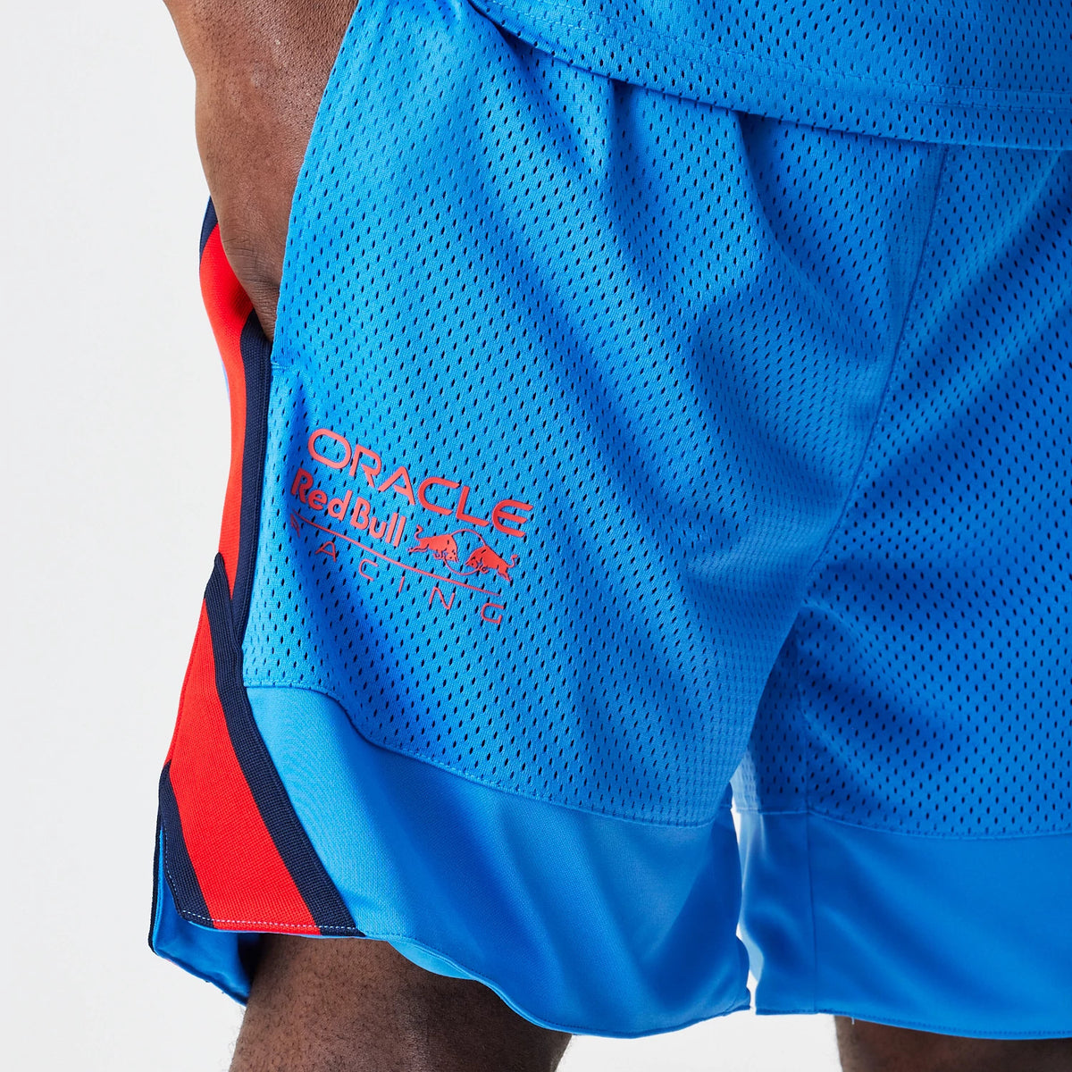 New Era Red Bull Racing Basketball Mesh Blue Shorts, front view showing a blue mesh fabric with a red Red Bull Racing logo printed on the left thigh, featuring a contrasting red and black side panel detail.