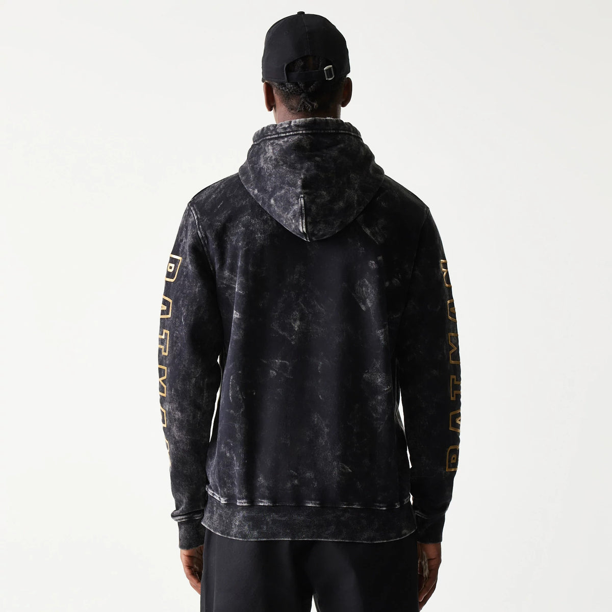 New Era Batman 1989 Black Pullover Hoodie, back view on a model with a black cap, showing a faded grey pattern across the hoodie and large Batman lettering in gold down each sleeve.