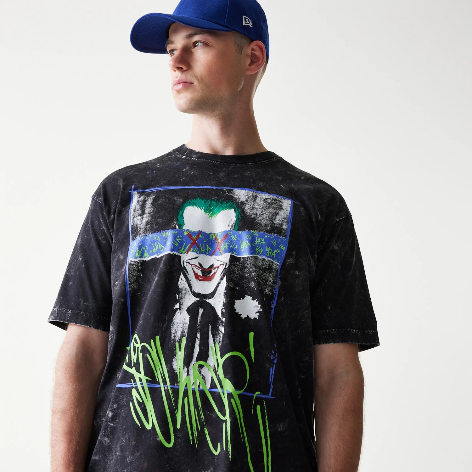 New Era Joker Batman Black T-Shirt, front view on a model wearing a blue cap, displaying a Joker graphic with green hair and red lips on the chest, and graffiti-style lettering in bright green at the bottom.
