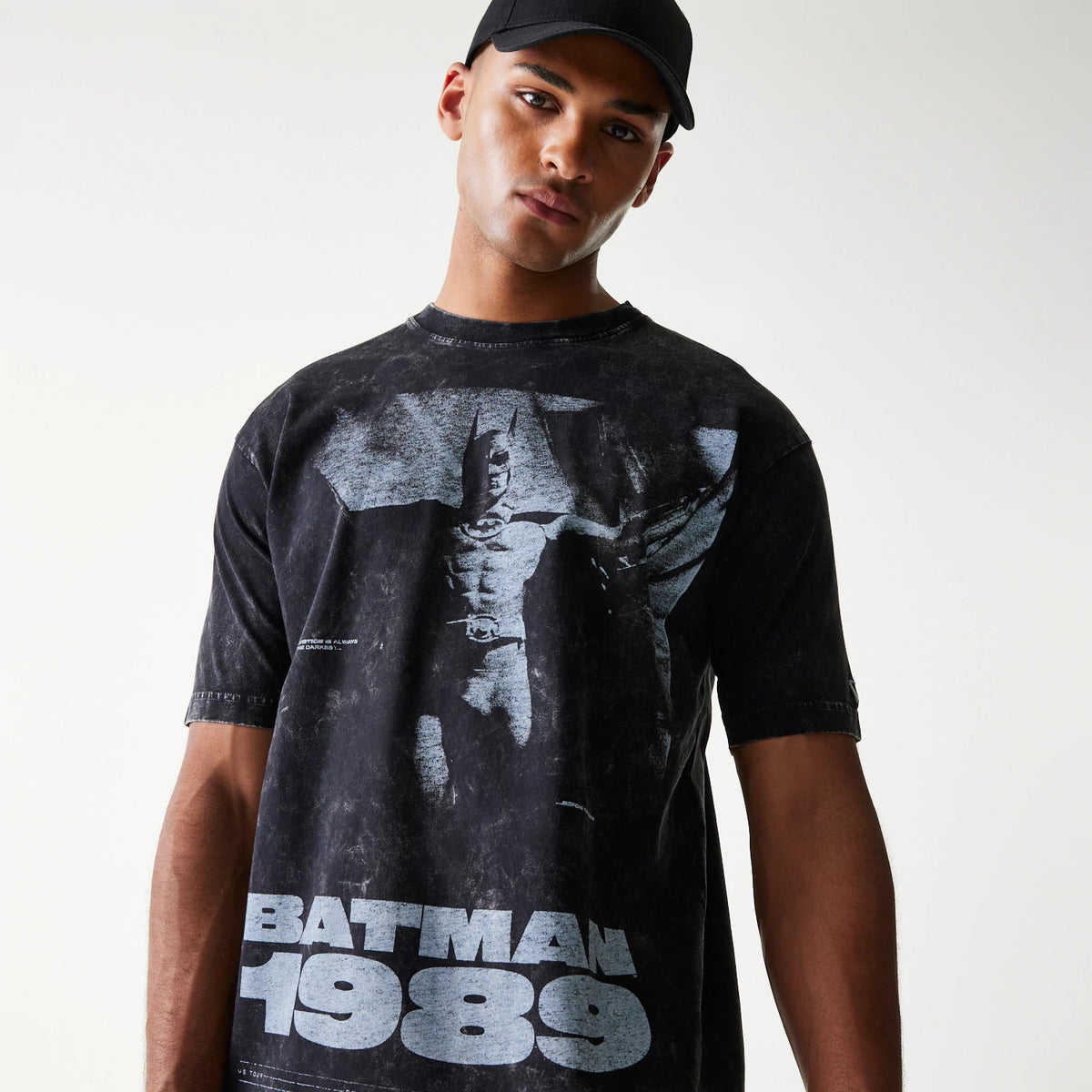 New Era Batman 1989 Black T-Shirt, front view on-model featuring a large graphic of Batman in grey tones across the chest with grey 'BATMAN 1989' lettering at the bottom, styled with a plain black cap.