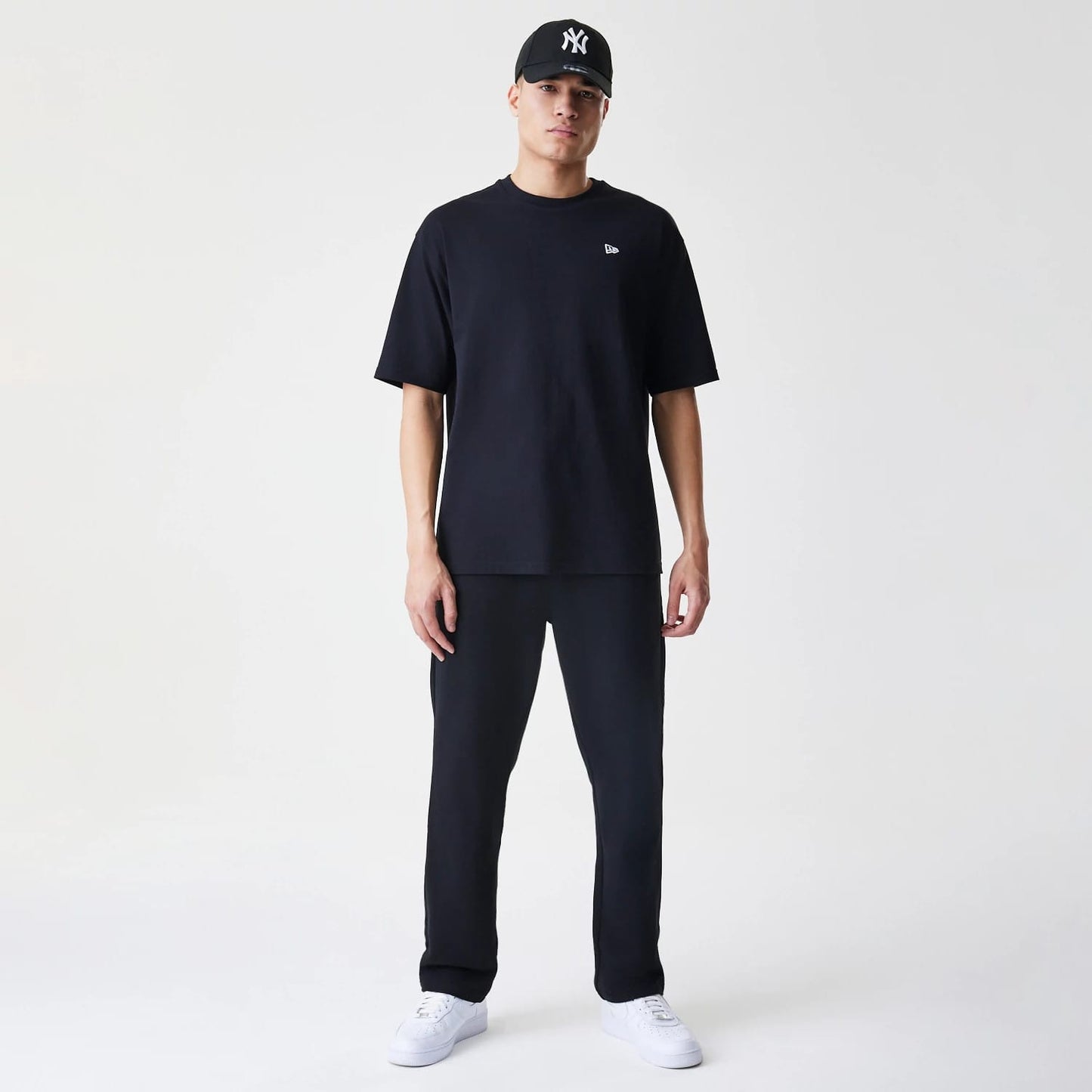 New Era Premium Essential Black Joggers, front view on a model styled with a matching black T-shirt and New York Yankees cap, featuring a relaxed fit and plain hems, paired with white trainers.