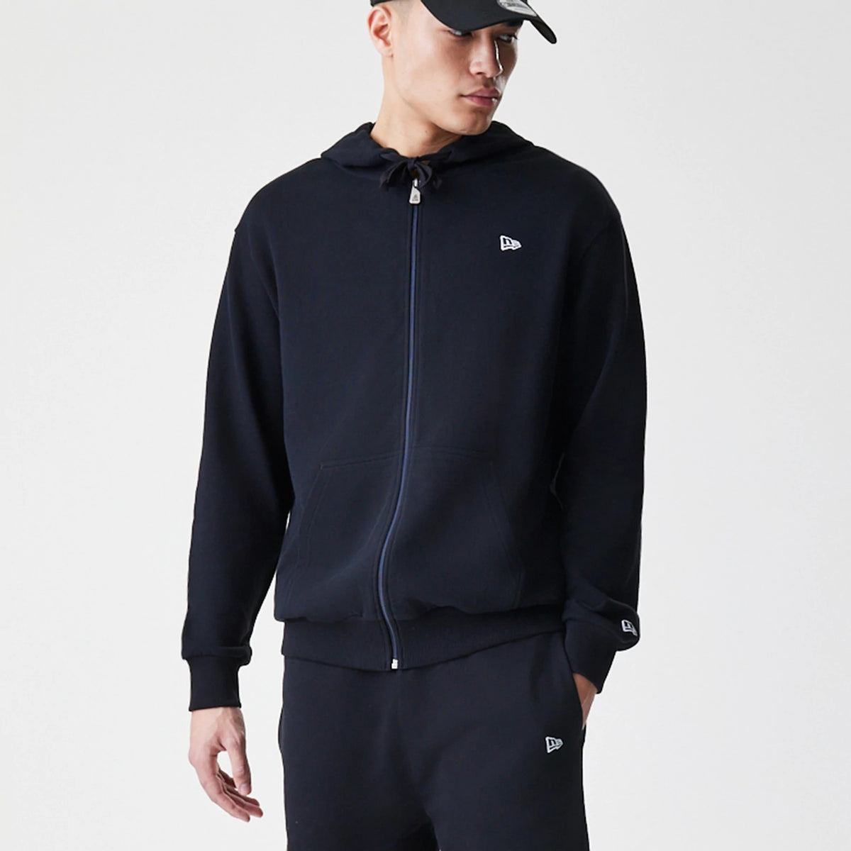 New Era Premium Essential Black Oversized Full Zip Hoodie, front view on-model showcasing a full-length zip, a drawstring hood, and an embroidered New Era logo on the chest and cuff, paired with matching black trousers and cap.