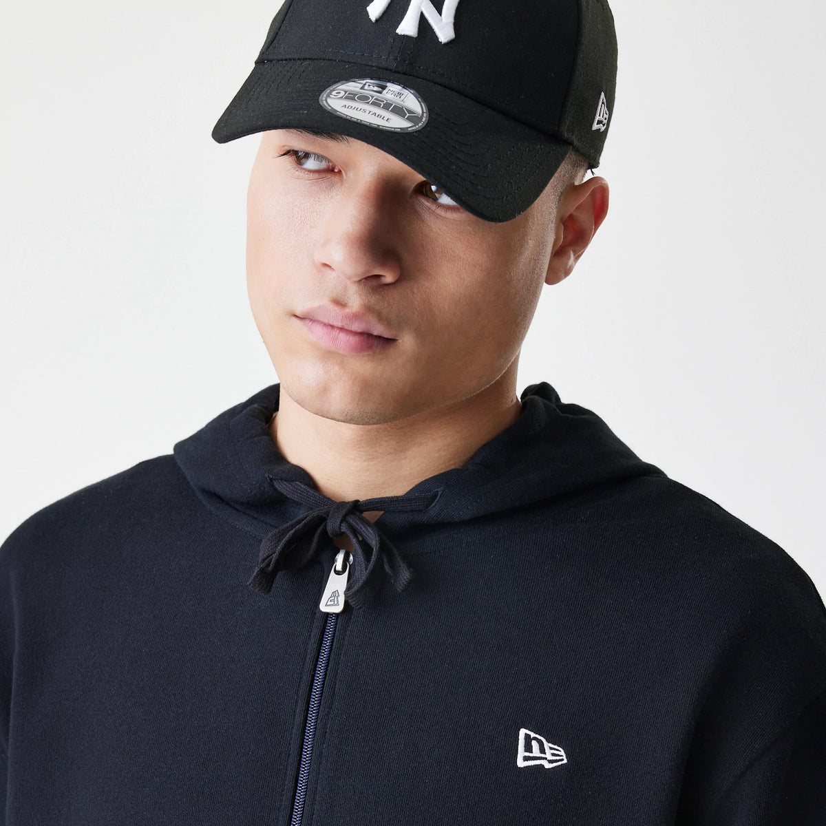 New Era Premium Essential Black Oversized Full Zip Hoodie, front view on-model showing the zip closure with a branded pull and an embroidered New Era logo on the left chest, styled beneath a black cap with logo details.