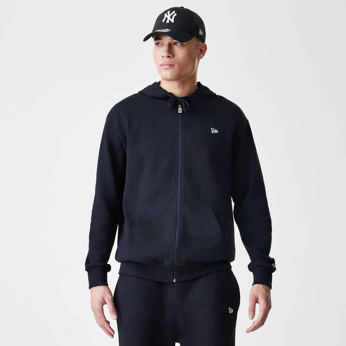 New Era Premium Essential Black Oversized Full Zip Hoodie, front view on a model showing the full zip closure, a embroidered New Era logo on the chest, and ribbed cuffs and hem paired with matching black trousers and cap.