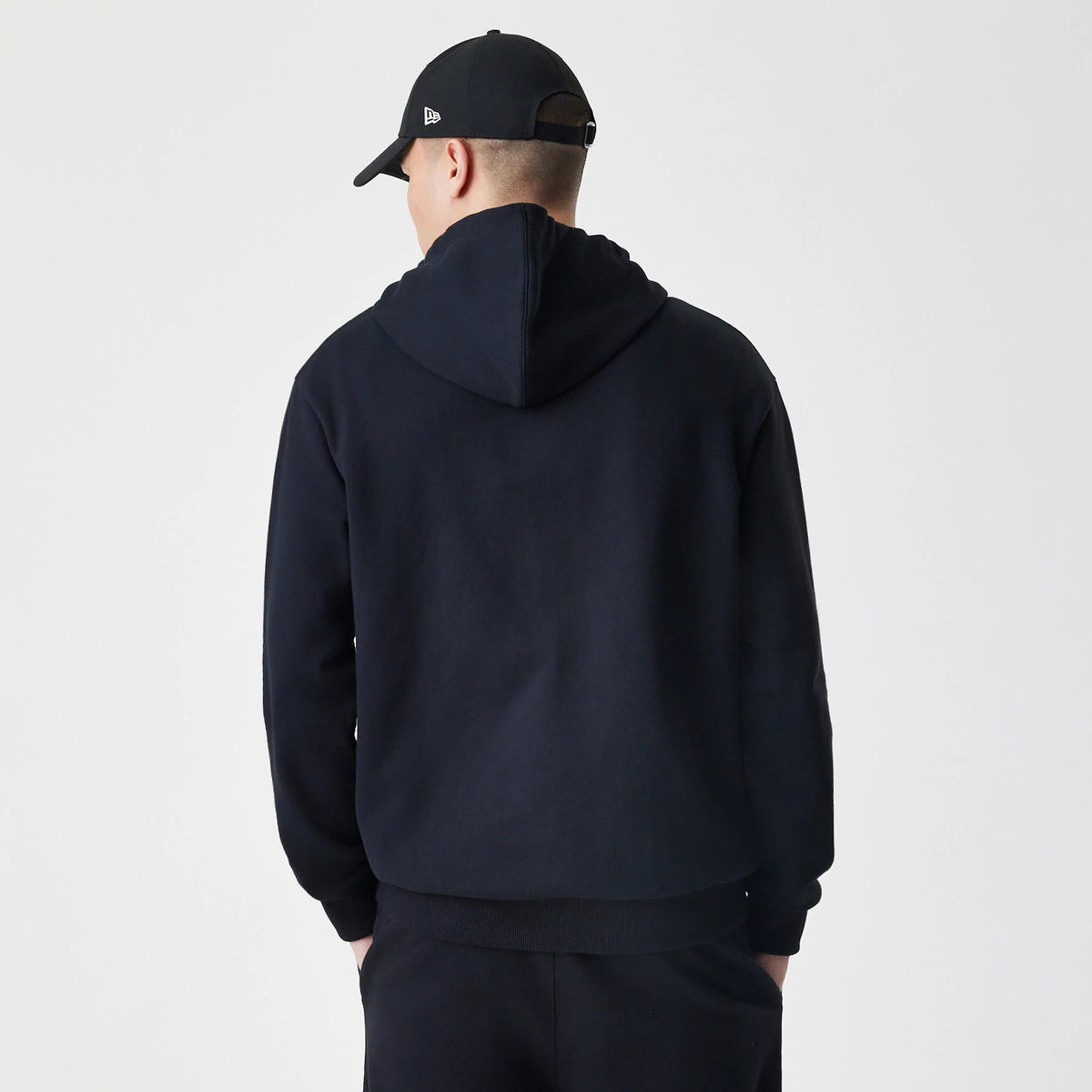 New Era Premium Essential Black Oversized Full Zip Hoodie, back view on a model with the hood down showing a plain black design and ribbed hem, paired with a black New Era cap featuring a white embroidered logo on the left side.