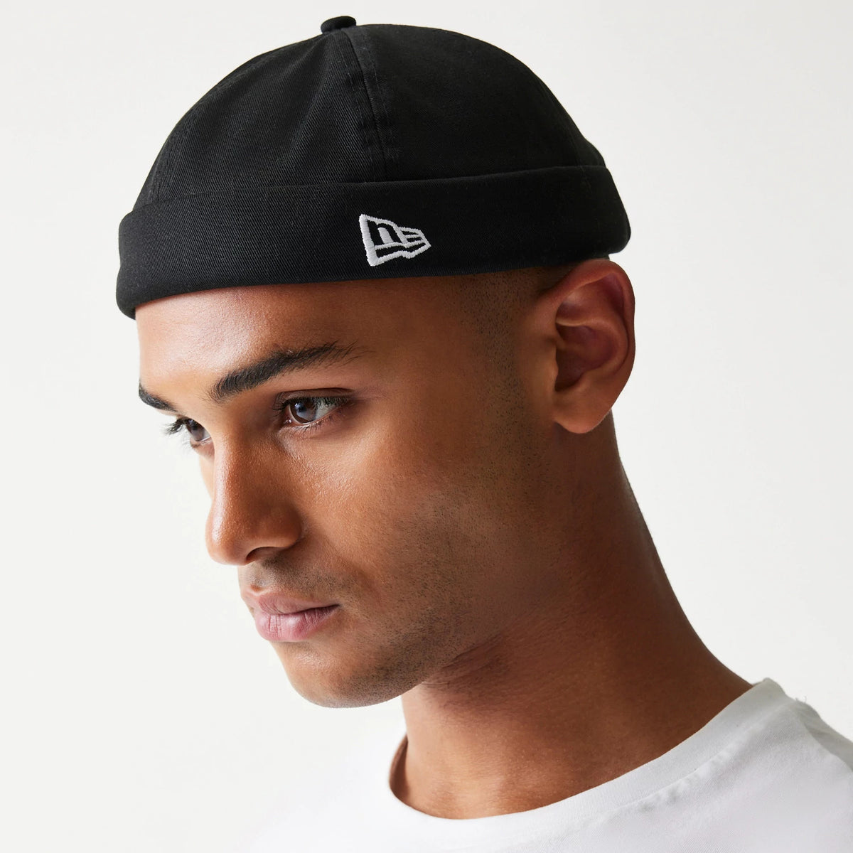New Era Cotton Twill Black Docker Hat, side view on a model with the white embroidered New Era flag logo on the turned-up cuff, showcasing the seamless round crown and black fabric.