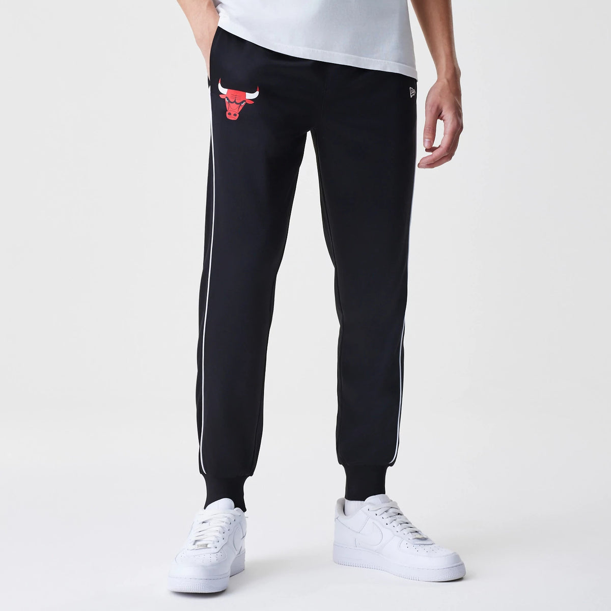 New Era Chicago Bulls NBA Slim Black Track Pants, front view on a model with a white shirt, featuring the red Bulls logo on the upper right leg, slim fit design with white piping down the sides and ribbed ankle cuffs.