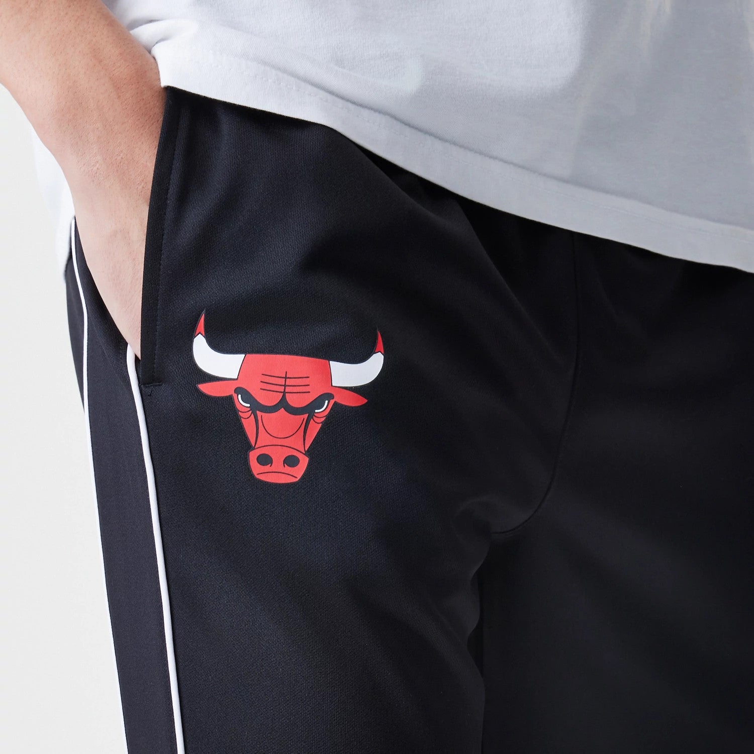 New Era Chicago Bulls NBA Slim Black Track Pants, close-up view showing the red Chicago Bulls logo on the black fabric near the left pocket, with white piping running down the seam for a contrast.