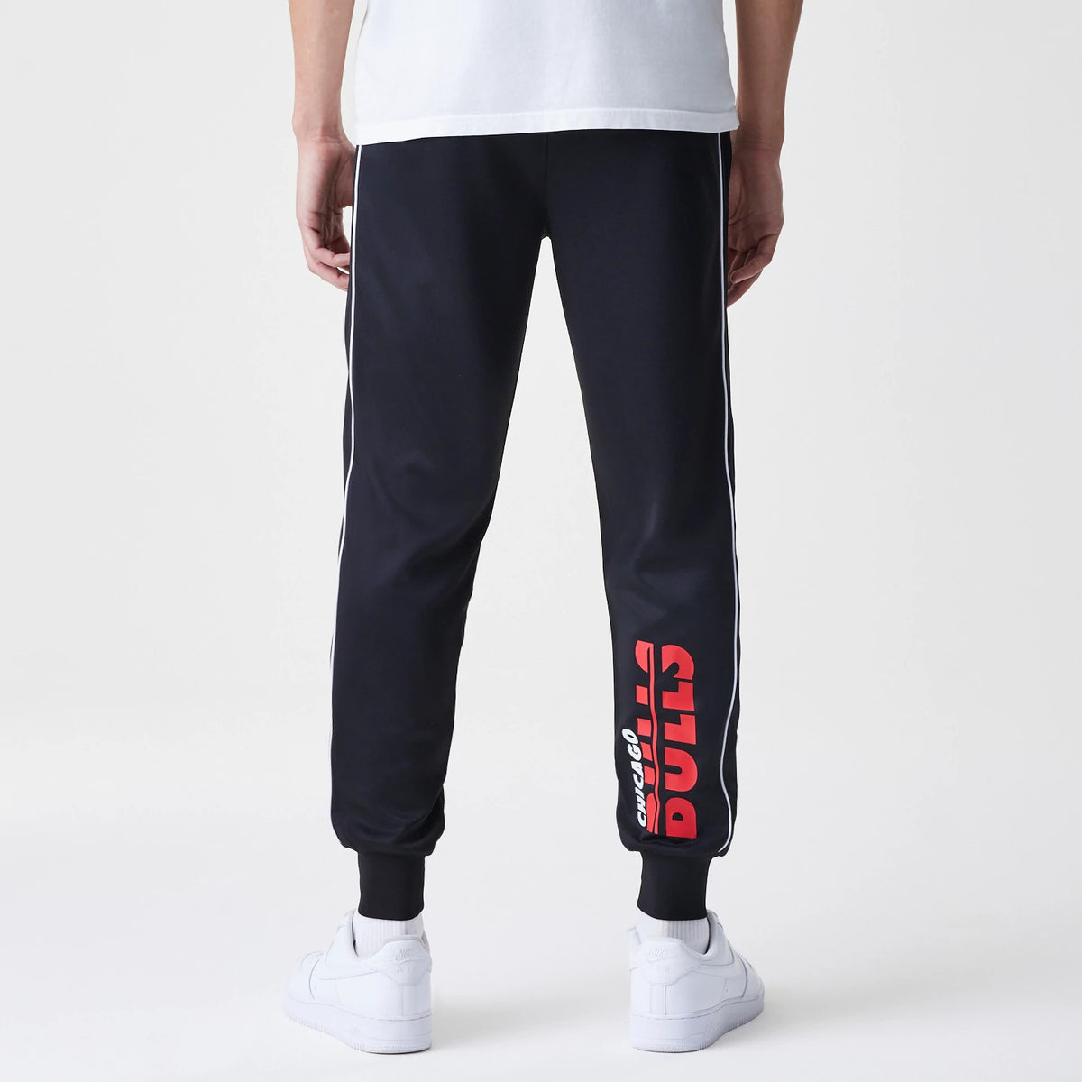 New Era Chicago Bulls NBA Slim Black Track Pants, rear view on a model showing red Chicago Bulls text down the left calf, with white piping along the sides and fitted black ankle cuffs.