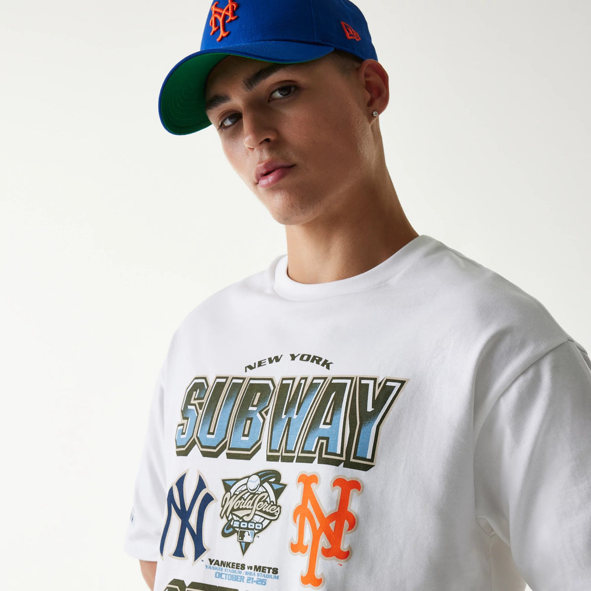 New Era MLB Subway Series White Oversized T-Shirt, front view on-model showing graphics of New York Yankees and Mets logos with 'Subway' text across the chest, styled with a matching blue cap.