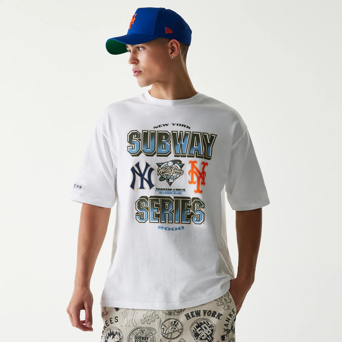 New Era MLB Subway Series White Oversized T-Shirt, front view on-model showing large printed graphics of New York Yankees and Mets logos with Subway Series lettering across the chest, styled with patterned shorts and a blue cap.