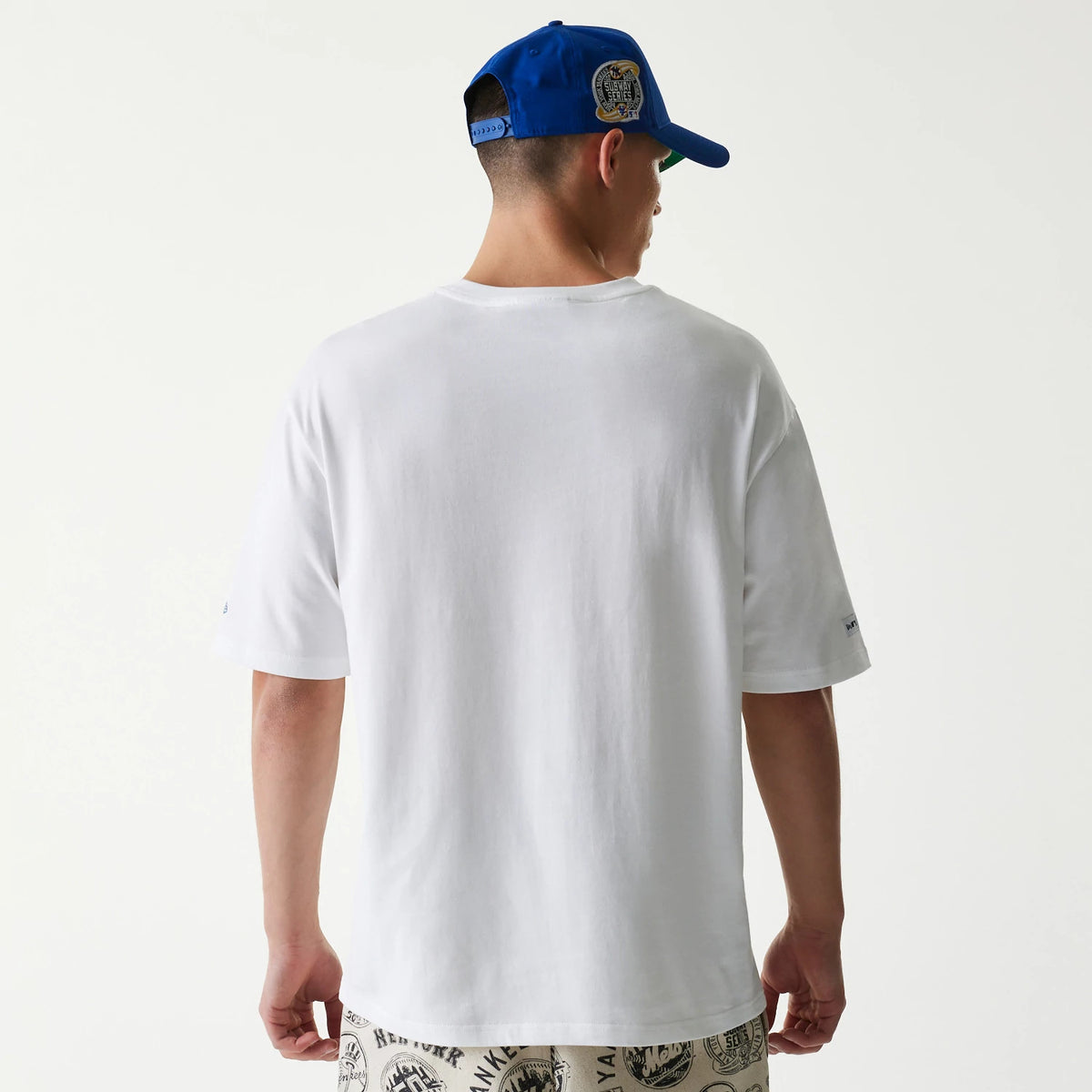 New Era MLB Subway Series White Oversized T-Shirt, back view on a model showing a plain white back with short sleeves, a embroidered New Era logo on the left sleeve and the shirt draping over printed shorts.