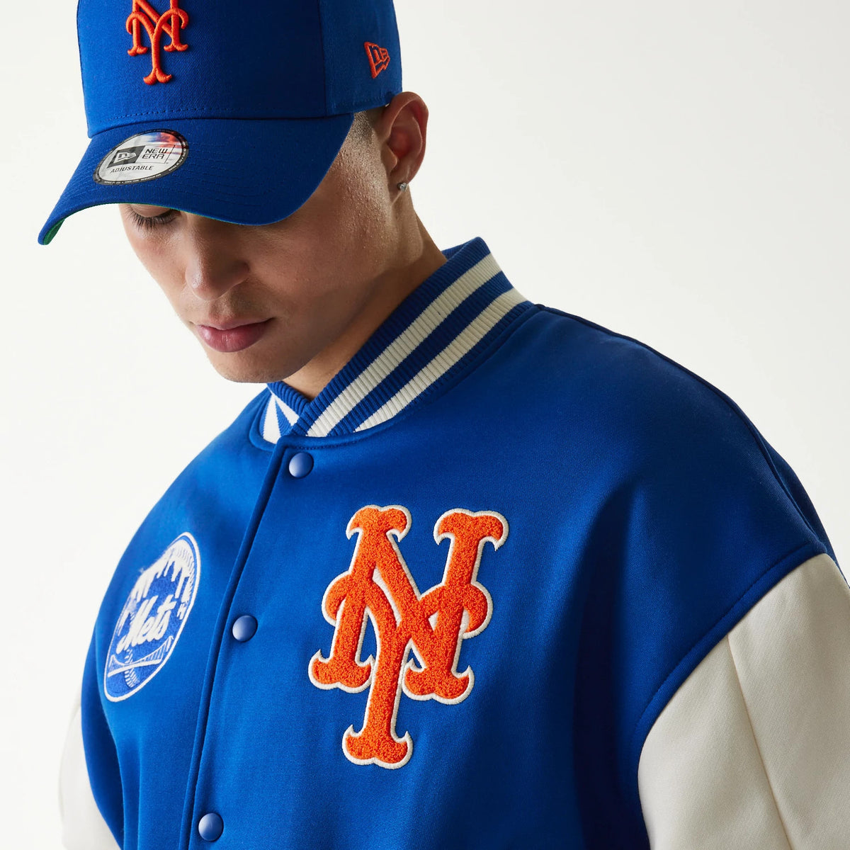 New Era New York Mets MLB Jersey Blue Bomber Jacket, front view showing an embroidered orange Mets logo on the chest, additional Mets circular patch, and striped blue and white ribbed collar, worn with a matching blue cap.