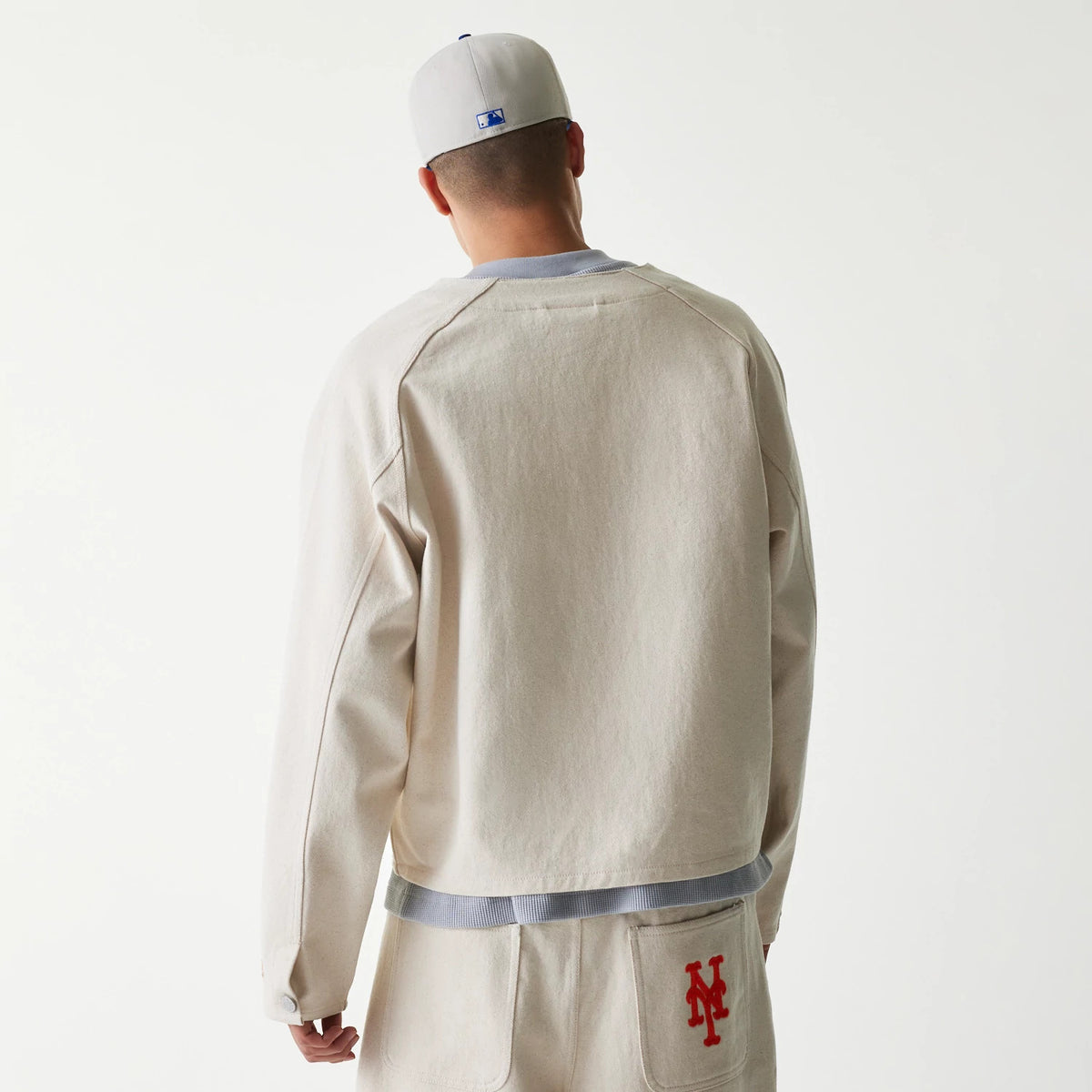 New Era New York Mets MLB Cotton Open Cream Jacket, back view on-model showing a plain back design and ribbed hem, styled with cream shorts featuring an embroidered red Mets logo on the back pocket.