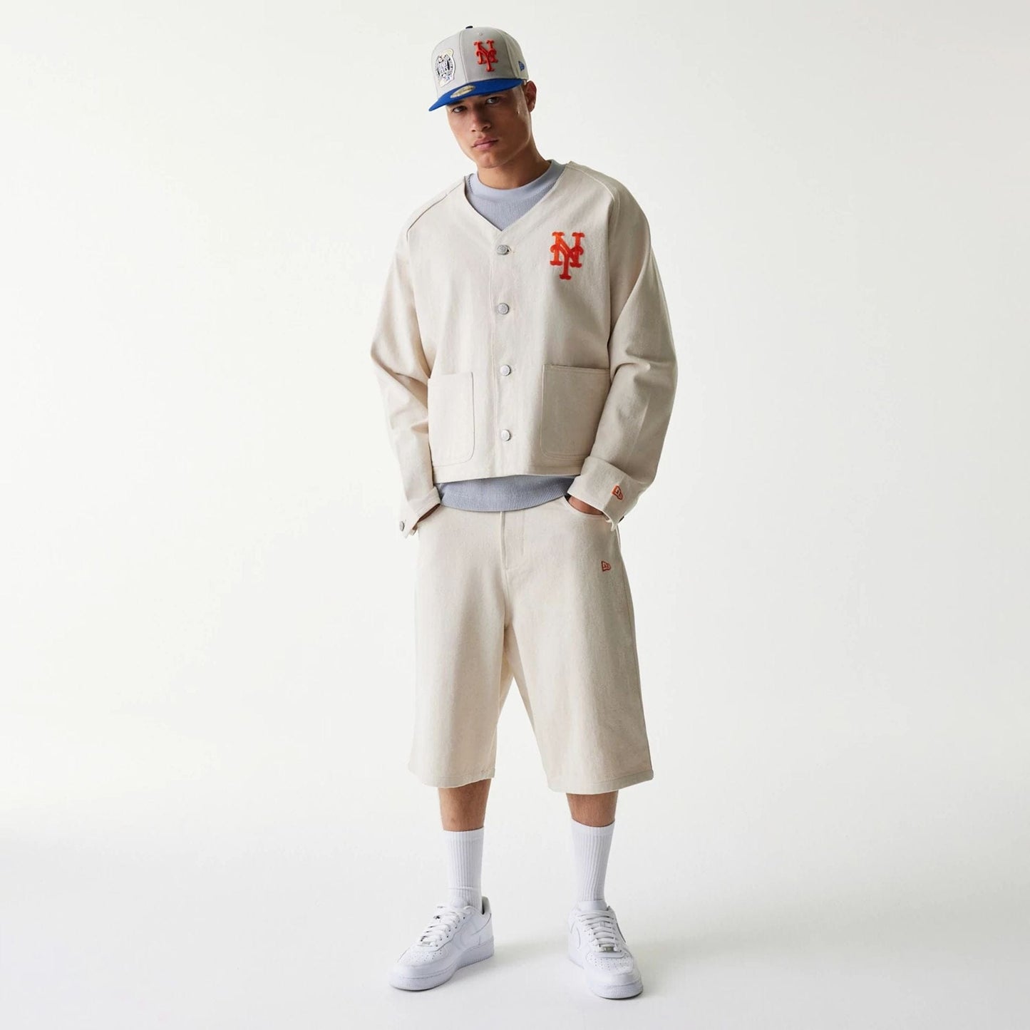 New Era New York Mets MLB Cotton Open Cream Jacket, front view on-model showing an orange embroidered Mets logo on the left chest, white buttons down the front, and worn with matching cream shorts and a cap.