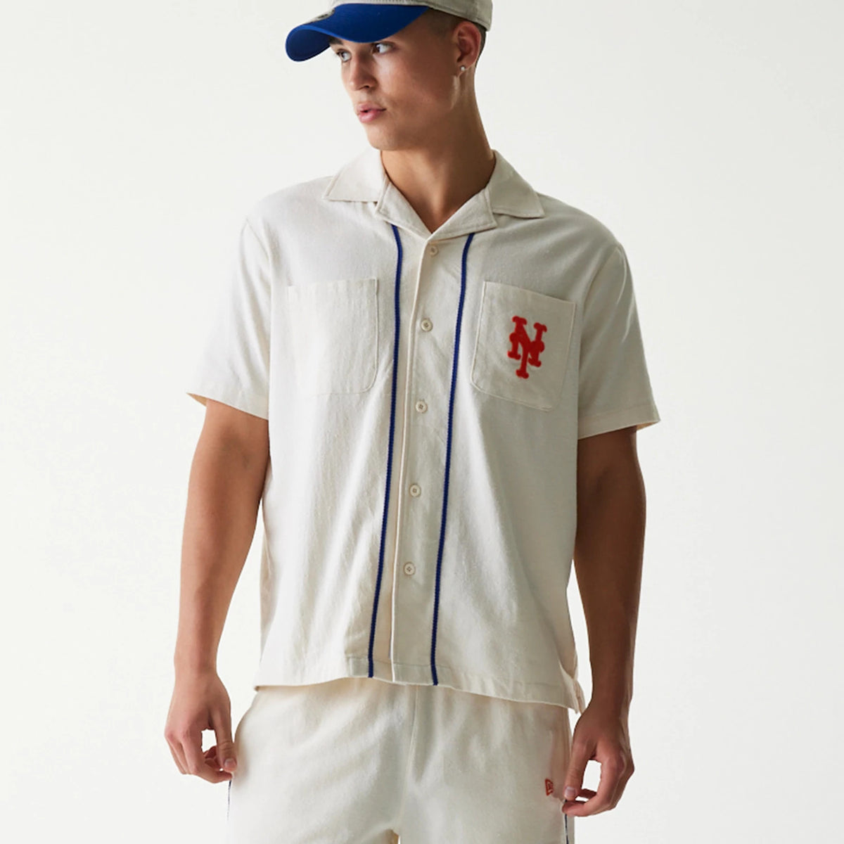 New Era New York Mets MLB Piping Open Cream Short Sleeve Shirt, front view on a model showing blue piping detail, button-down design, and red embroidered NY logo on the left chest pocket.