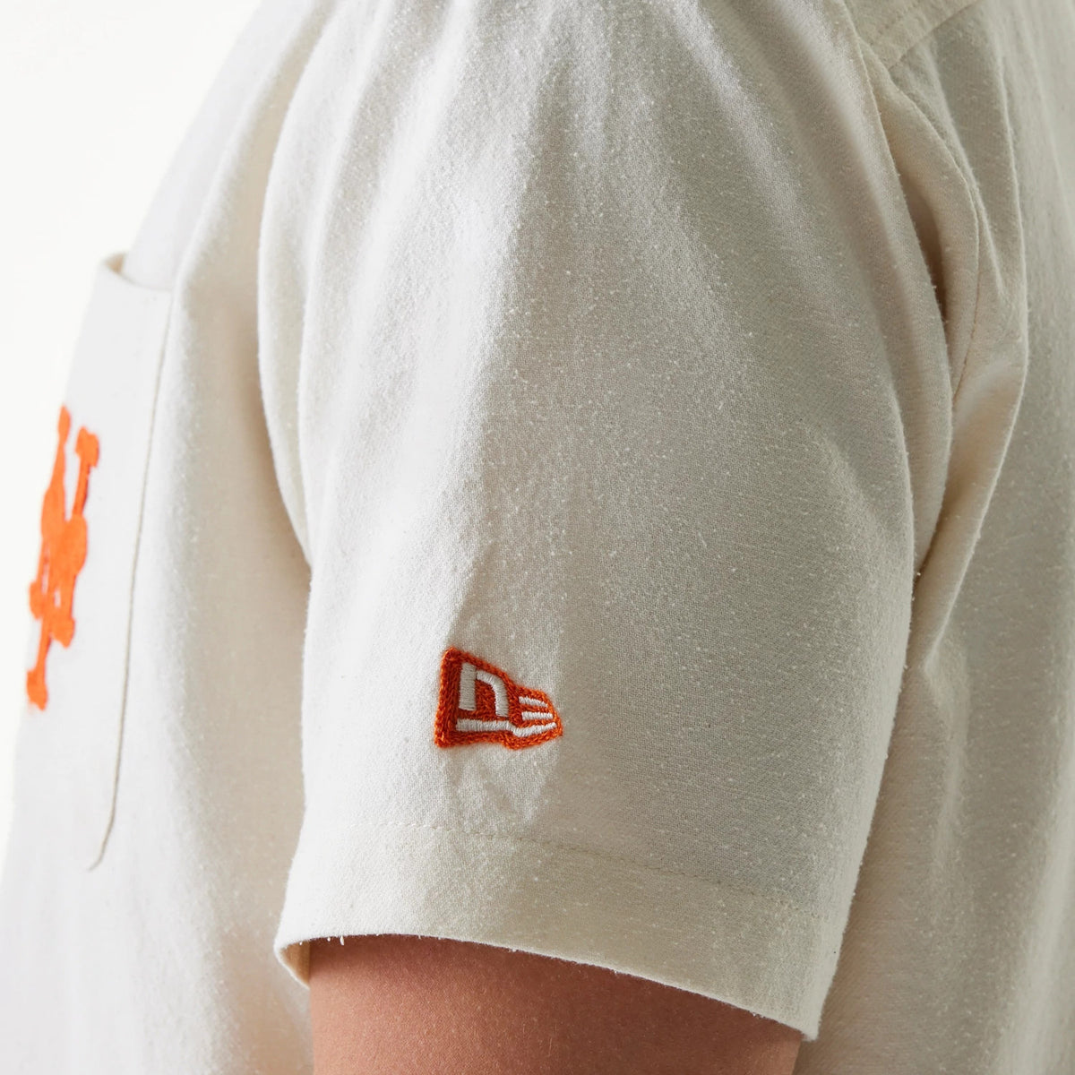New Era New York Mets MLB Piping Open Cream Short Sleeve Shirt, side view showing the orange New Era flag logo embroidered on the sleeve and the edge of the orange Mets logo on the chest.