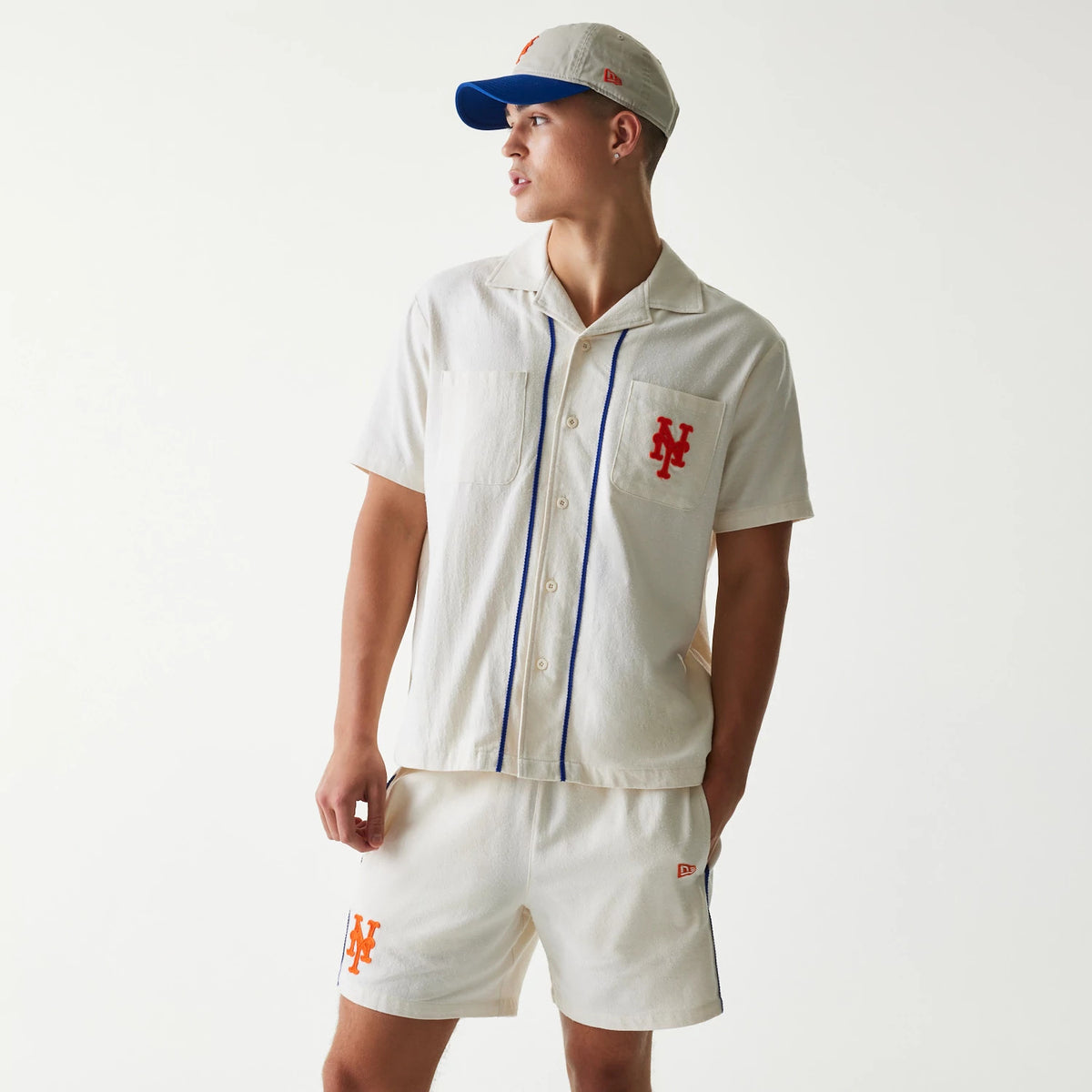 New Era New York Mets MLB Piping Open Cream Short Sleeve Shirt, front view on a model with a matching cap, featuring blue piping along the button placket and red Mets logo embroidered on the left chest pocket.