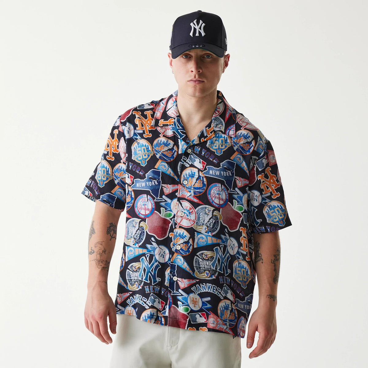 New Era MLB Subway Series All Over Print Navy Short Sleeve Shirt, front view on a model wearing a matching cap, featuring a mix of New York Yankees and Mets logos in orange, blue and white across the entire navy fabric.