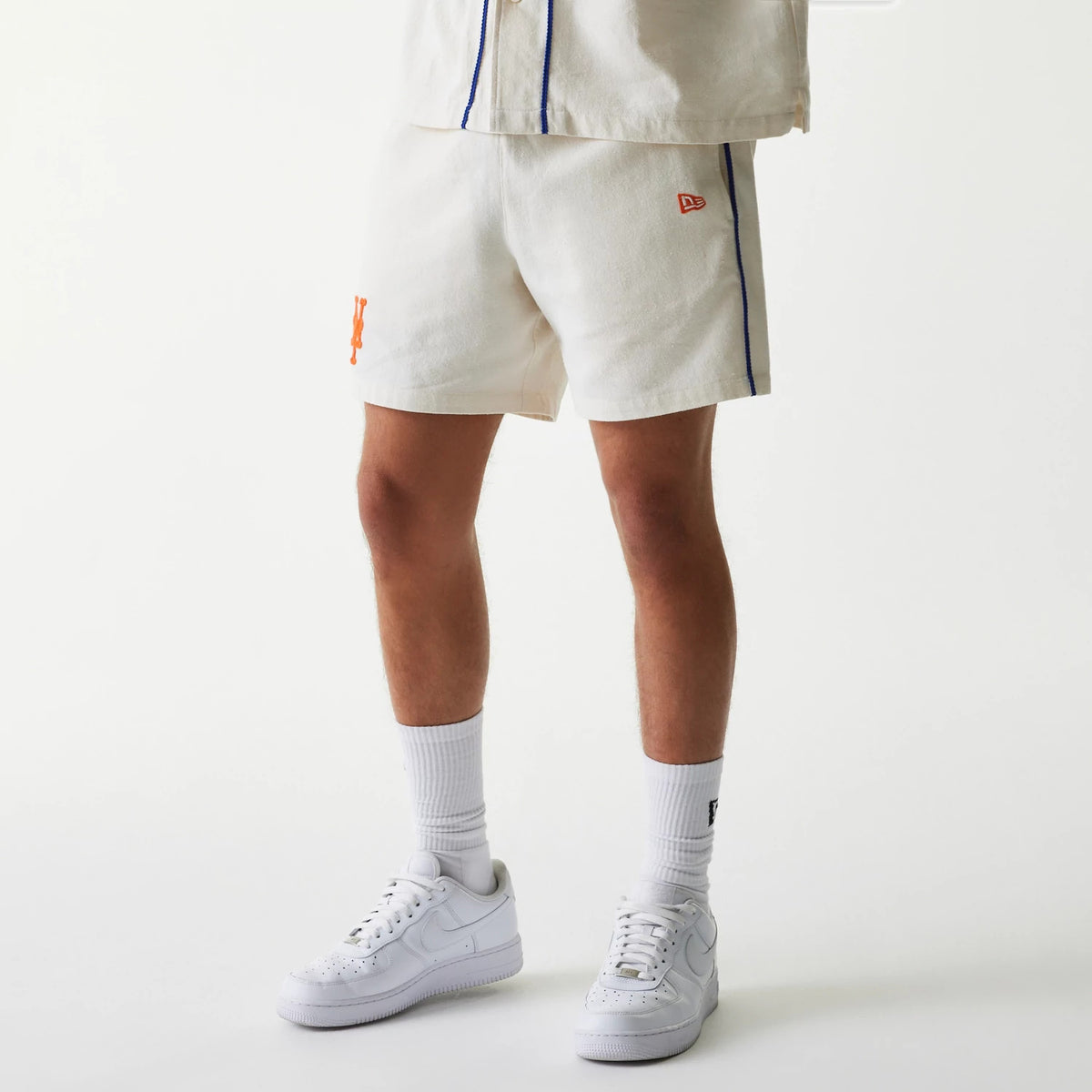 New Era New York Mets MLB Piping Open Cream Shorts, front view showing the cream shorts with blue piping along the side, an orange embroidered Mets logo near the hem, and a New Era logo above the left hem, styled with white trainers and socks.