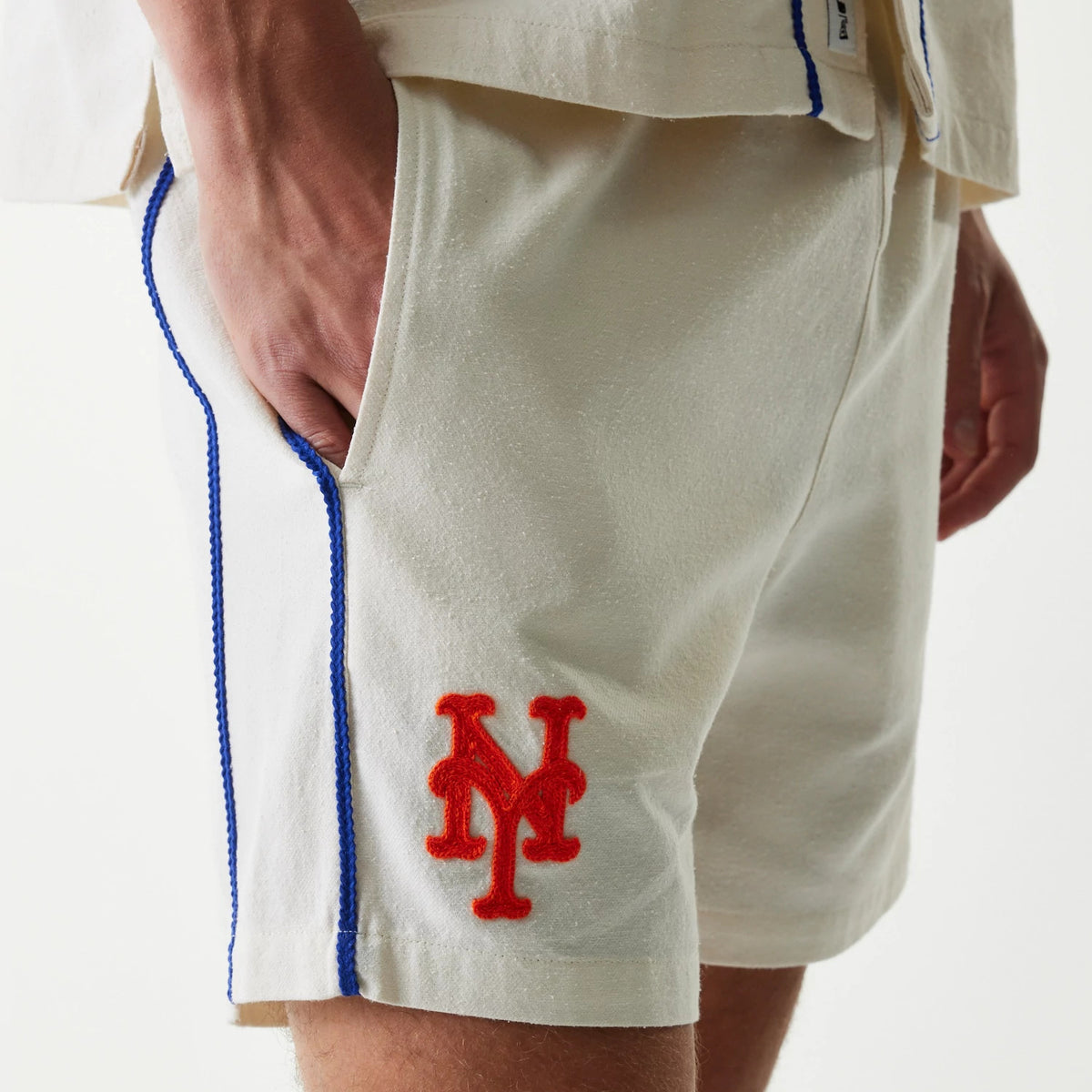 New Era New York Mets MLB Piping Open Cream Shorts, three-quarter view showing the cream shorts with a bright red embroidered NY logo on the left leg and blue piping detail running along the side seam.