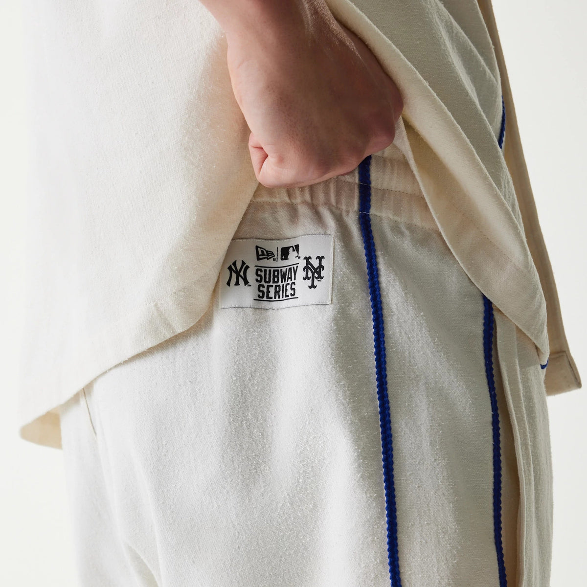 New Era New York Mets MLB Piping Open Cream Shorts, side view showing the cream fabric with blue piping running vertically and a Subway Series patch printed on the waistband, styled with a matching cream top.