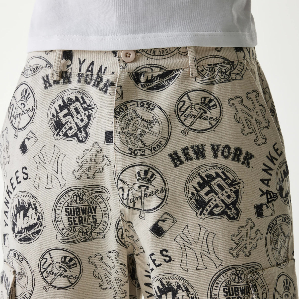 New Era MLB Subway Series All Over Print Cargo Trousers, close-up view showing a beige fabric with black logos and emblems of the New York Yankees and Mets, button fastening, belt loops and detailed stitching near the pockets.