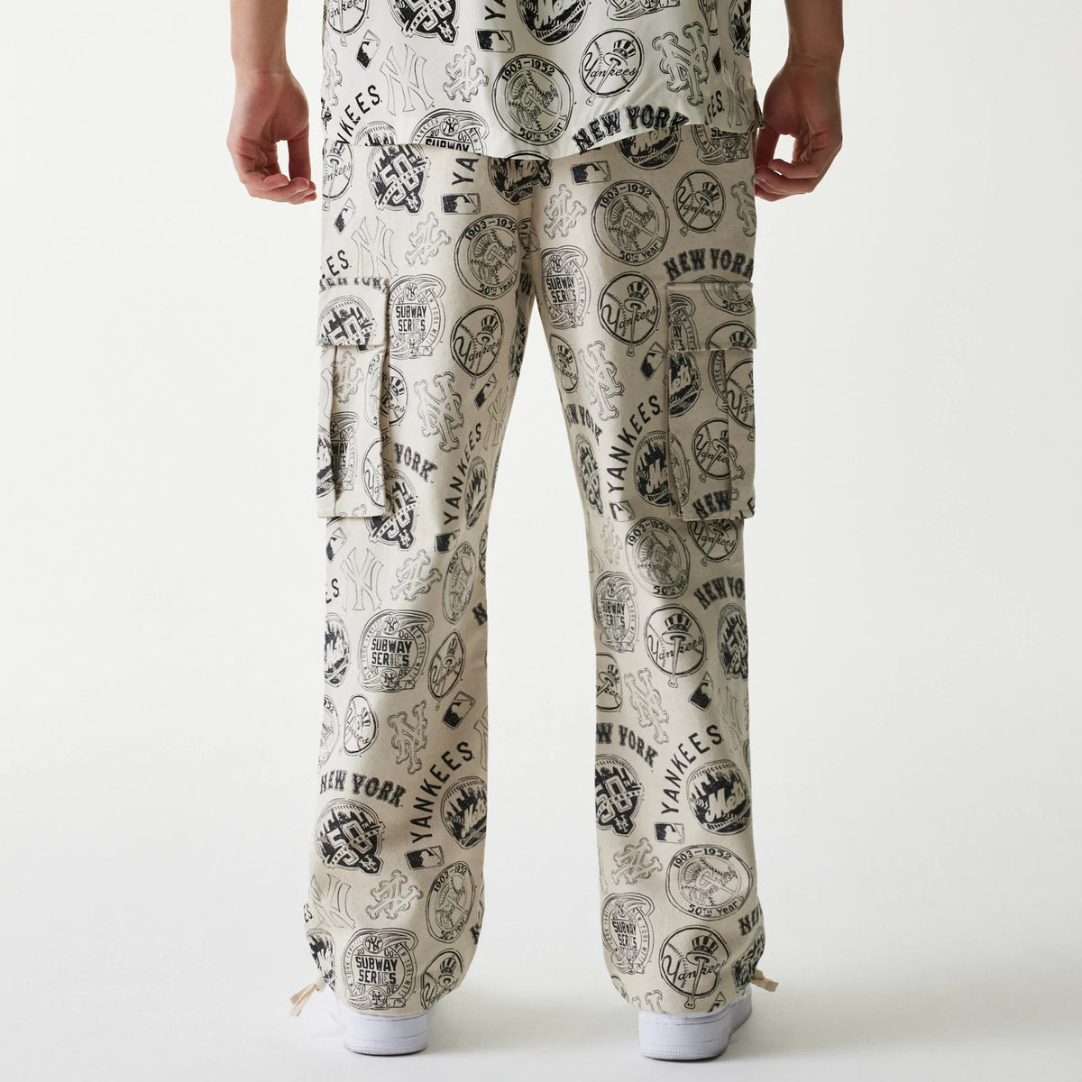 New Era MLB Subway Series All Over Print Cargo Trousers, rear view on a model showing the wide-leg fit with multiple New York Yankees themed graphics, including logos and signatures, printed across the cream fabric.
