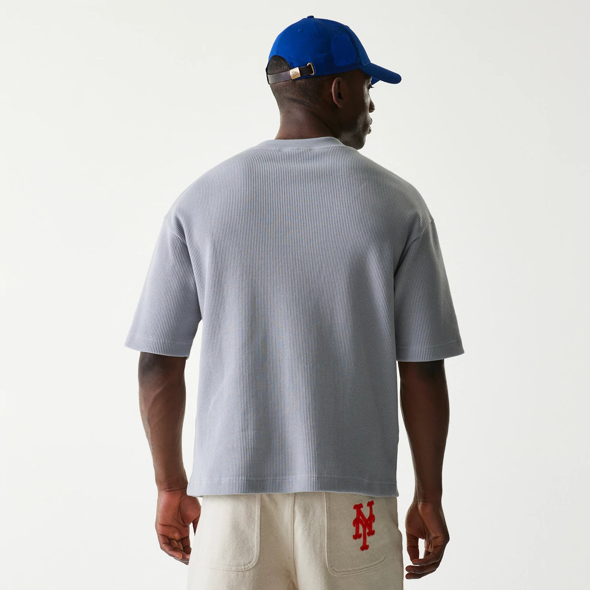 New Era New York Mets MLB Waffle Grey Oversized T-Shirt, back view on a model featuring a simple ribbed texture, complemented by neutral shorts with a red embroidered team logo on the pocket and a blue cap.