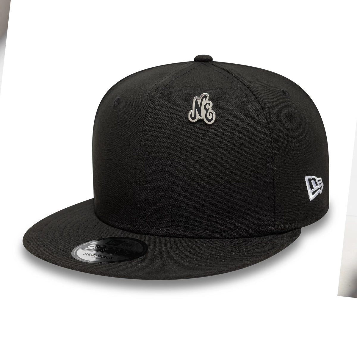 New Era Script Black Pin Badge, three-quarter view showing a pin badge with the initials NE on the front crown of a black cap, a white embroidered New Era logo on the side, and a flat black visor with a sticker.