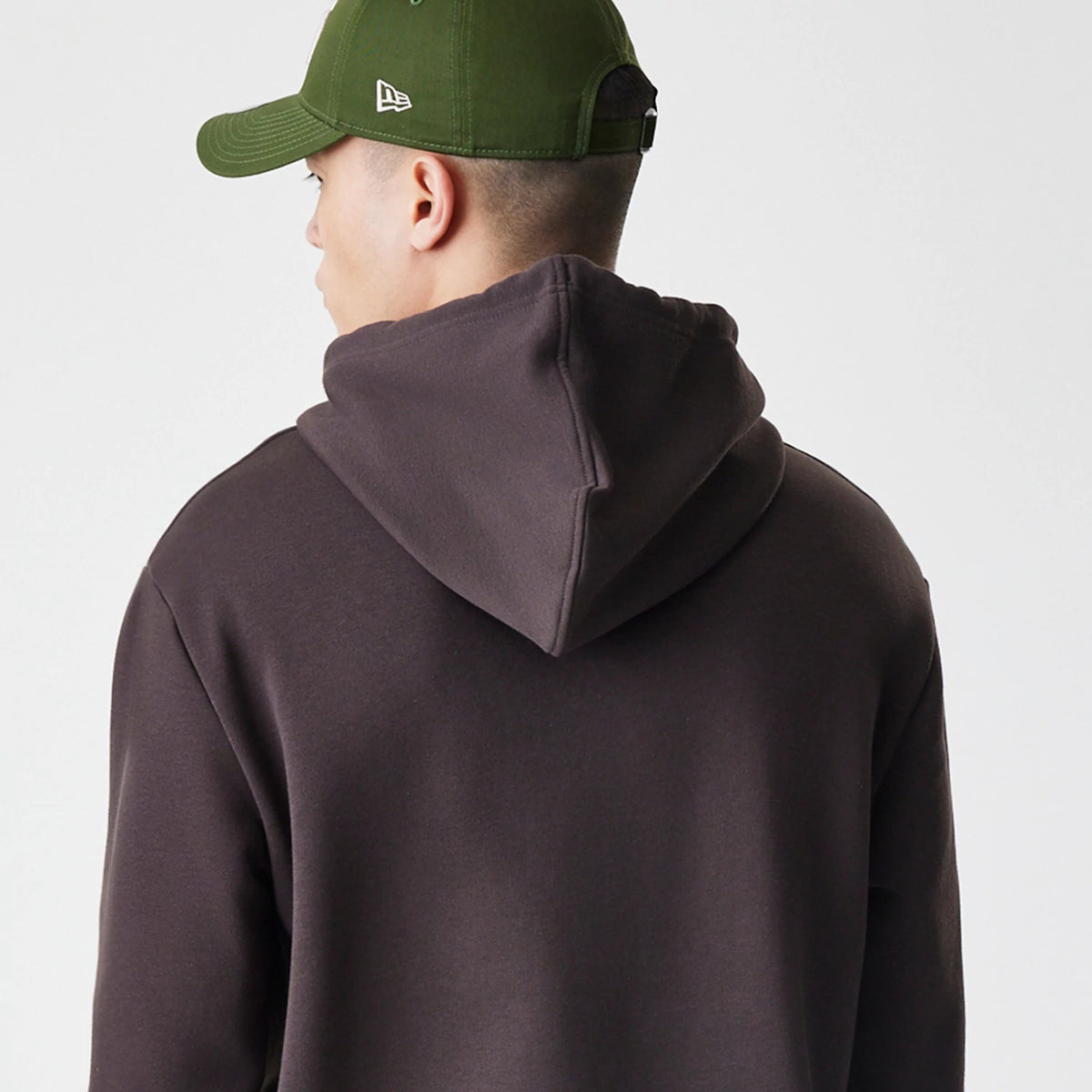 New Era New York Yankees MLB Midi League Essential Dark Brown Oversized Pullover Hoodie, back view on-model showing the plain dark brown hood with its structure, styled with a green cap featuring an embroidered New Era logo on the side.