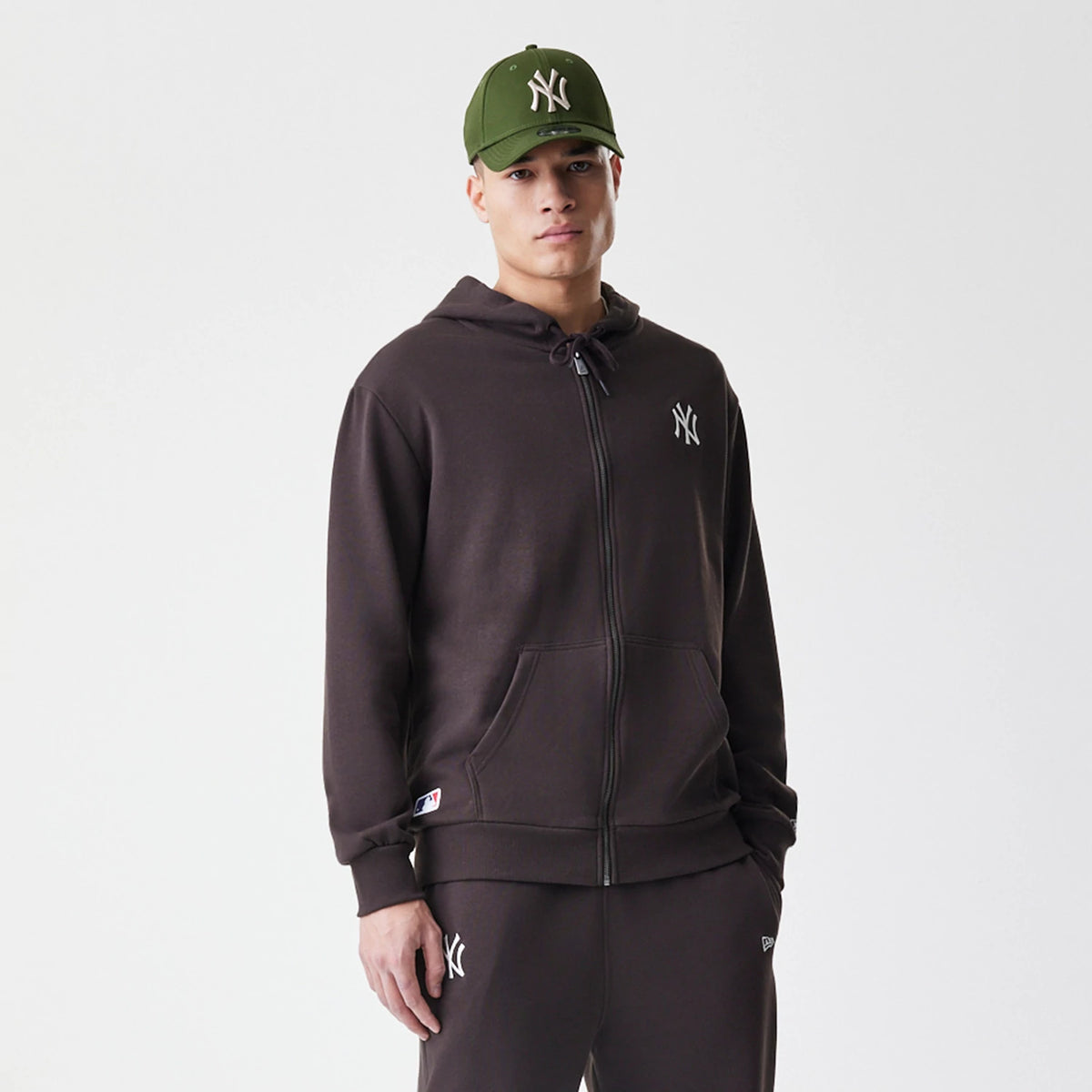 New Era New York Yankees MLB Midi League Essential Dark Brown Full Zip Hoodie, front view on-model showing the white embroidered NY logo on the left chest, a full-length zip, and drawstring hood, styled with matching dark brown joggers.