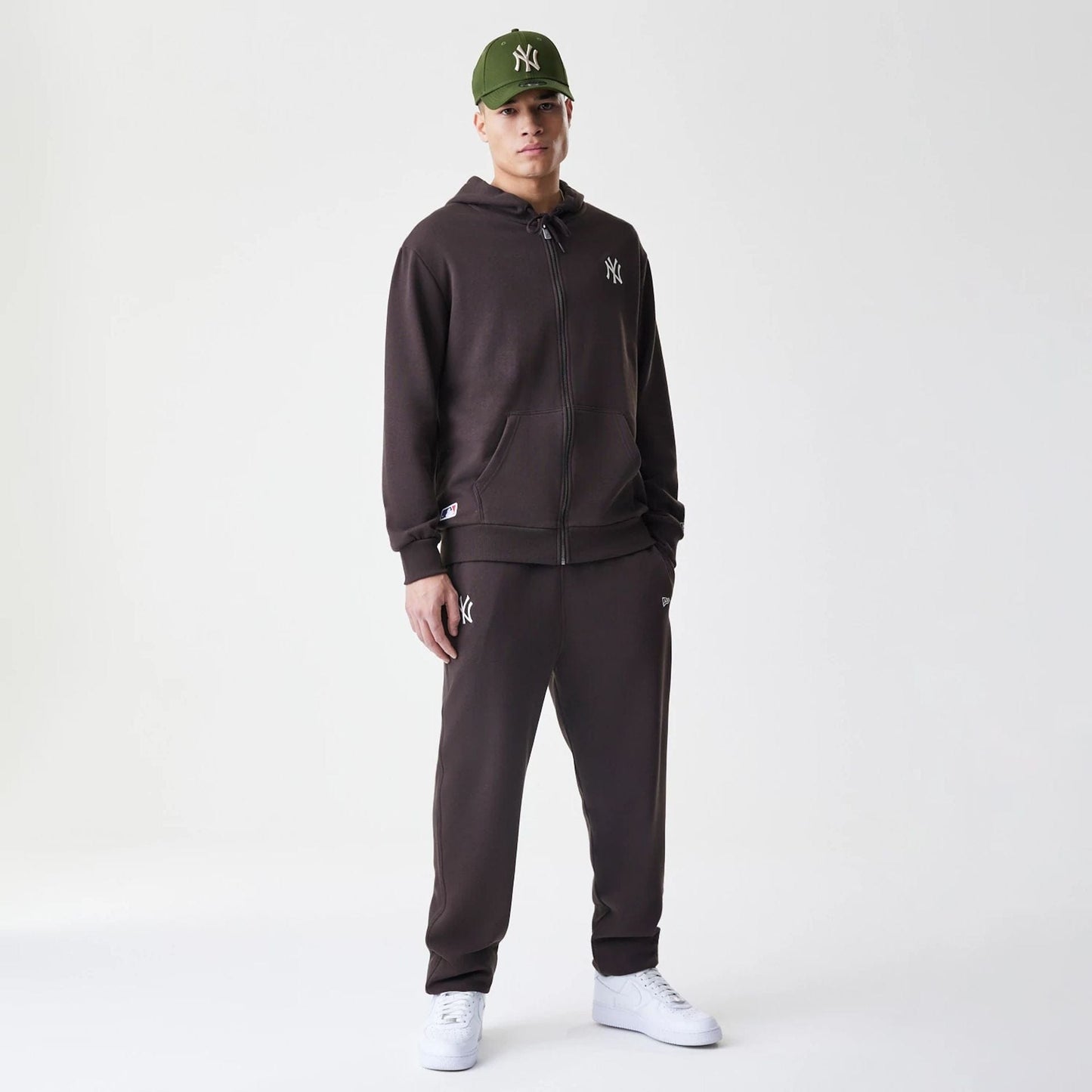 New Era New York Yankees MLB Midi League Essential Dark Brown Full Zip Hoodie, front view on-model showing the dark brown hoodie with an embroidered white NY logo on the left chest, paired with matching trousers and a green cap.