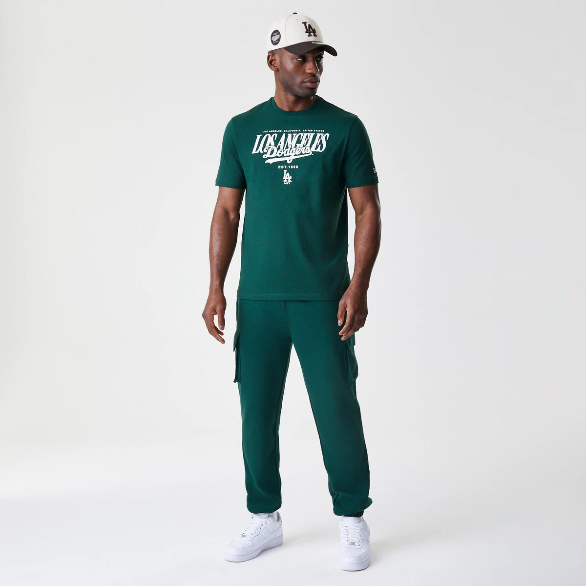 New Era LA Dodgers MLB Script Graphic Dark Green T-Shirt, front view on-model showing the white Los Angeles Dodgers script across the chest, with a small embroidered New Era logo near the sleeve hem.